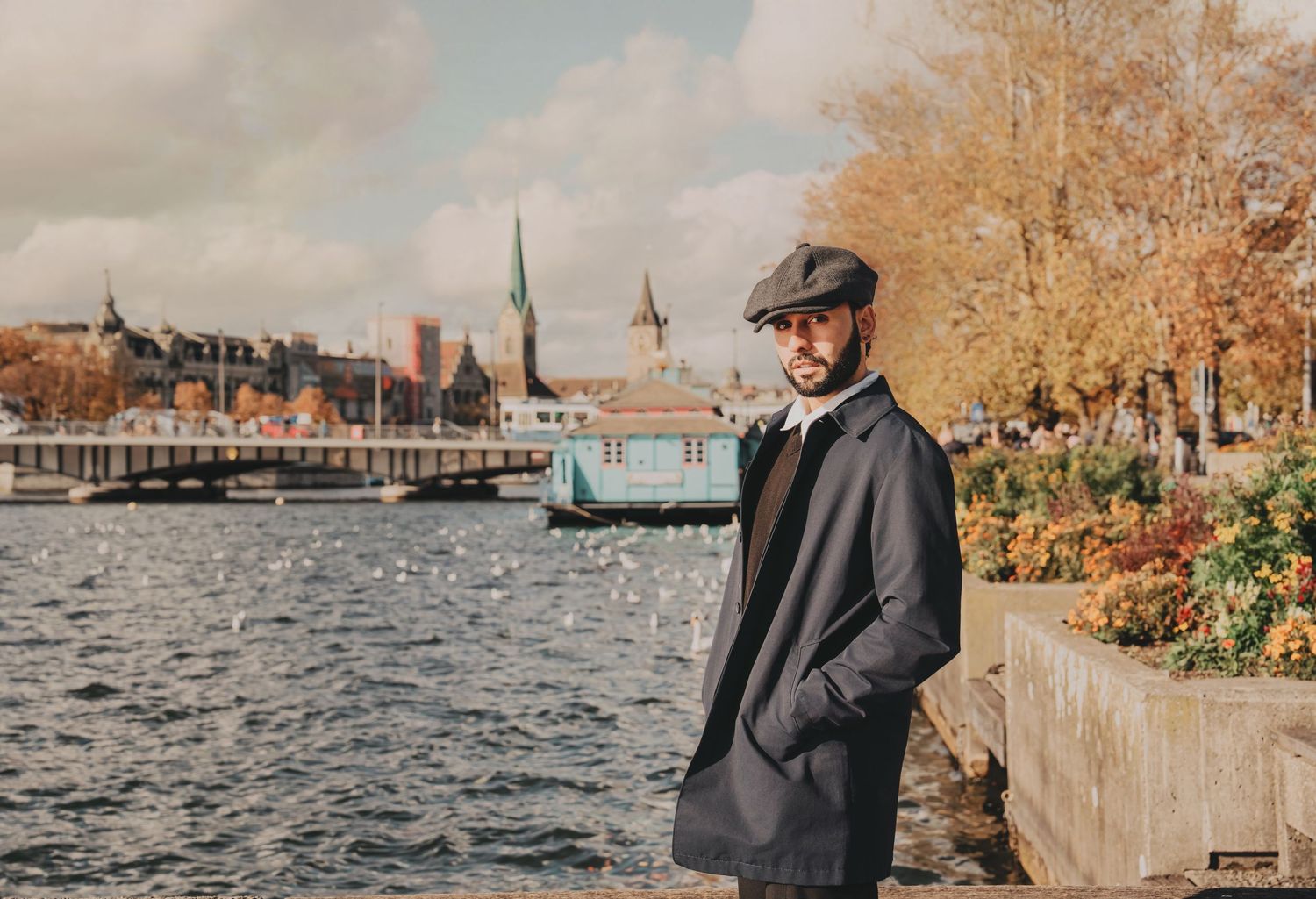 Stylish man in black coat and flat cap standing by waterfront in autumn cityscape.