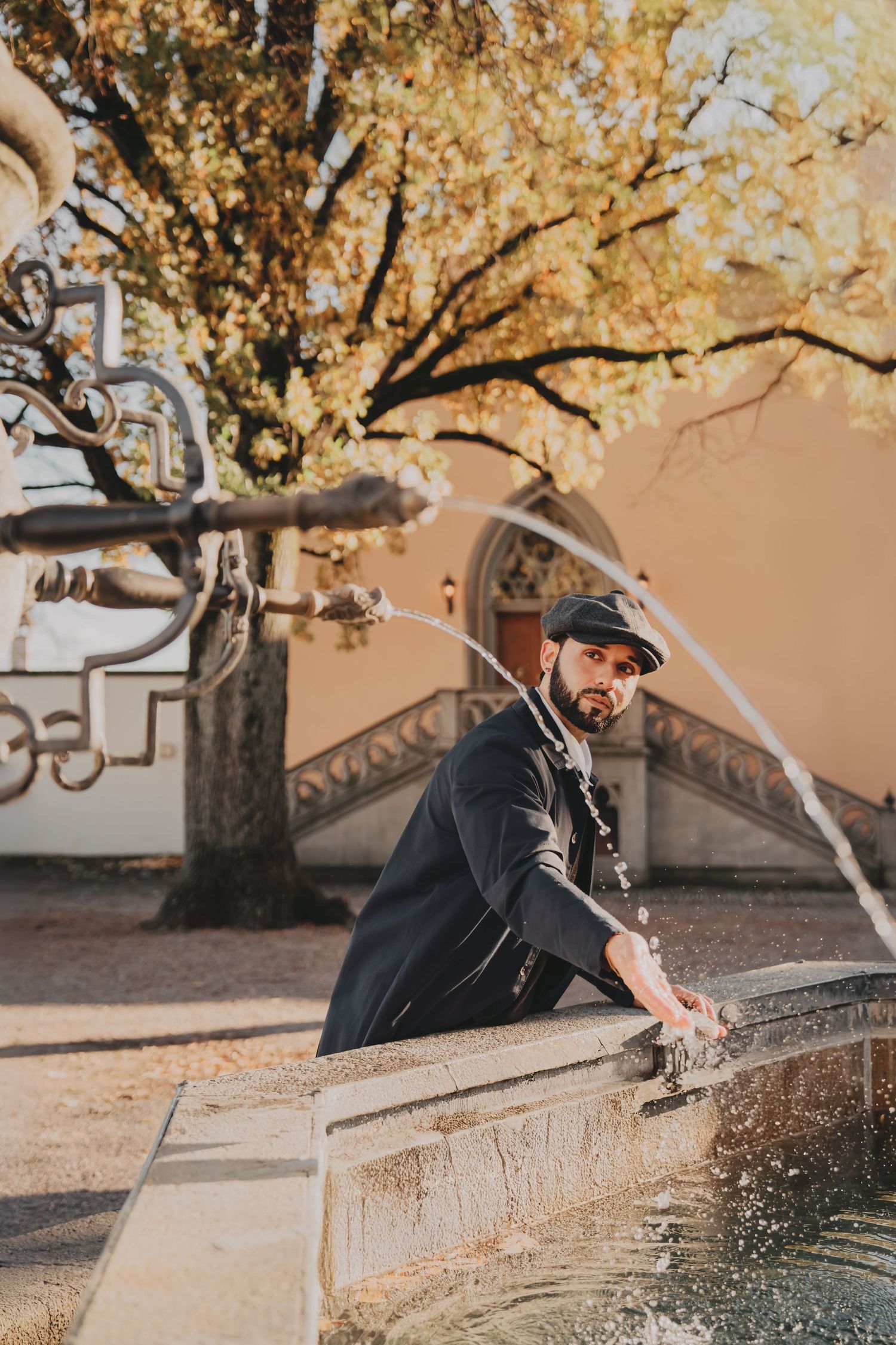 Bearded man in black coat and flat cap leaning over a decorative stone fountain with autumn trees.
