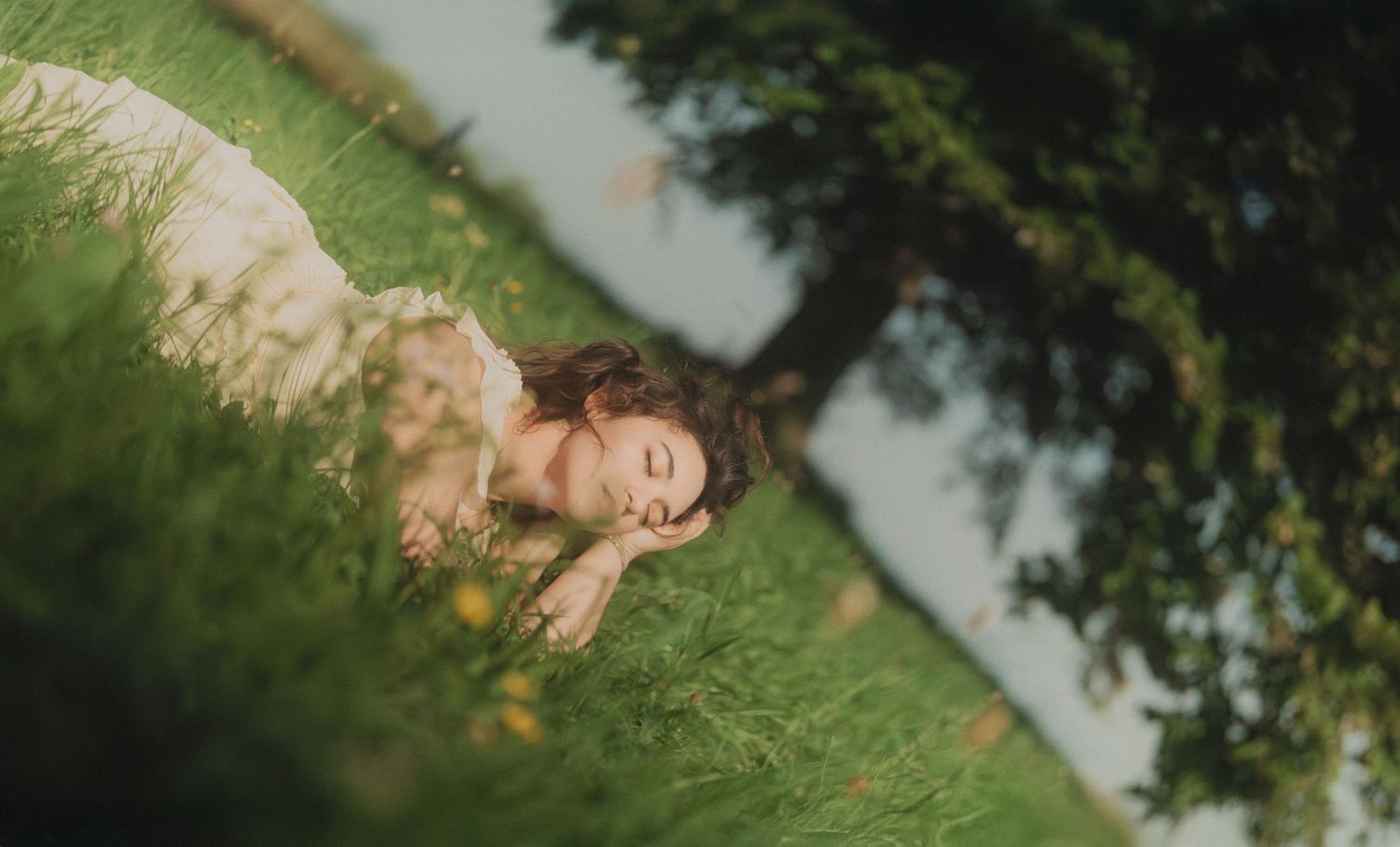 Aerial view of woman in white dress lying peacefully in lush green grass beneath a tree.