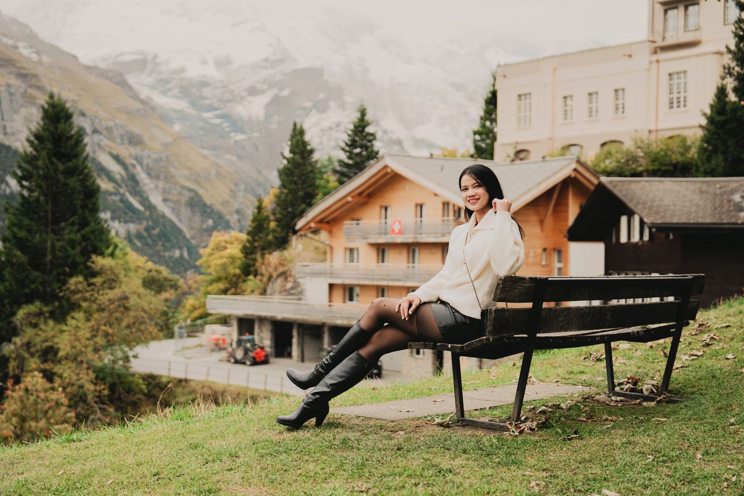 Smiling Asian woman in white sweater sits on park bench with Swiss chalet and misty mountains behind her.