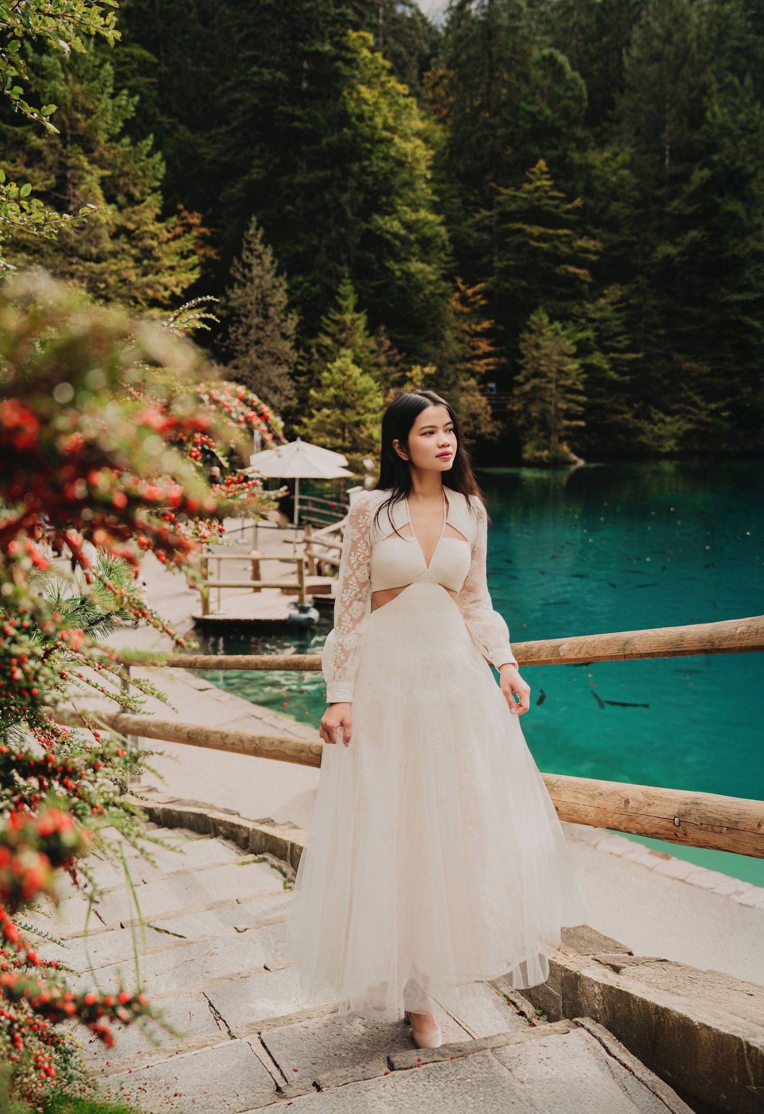 Woman in white lace dress descends stone steps beside vivid turquoise lake and red berries.