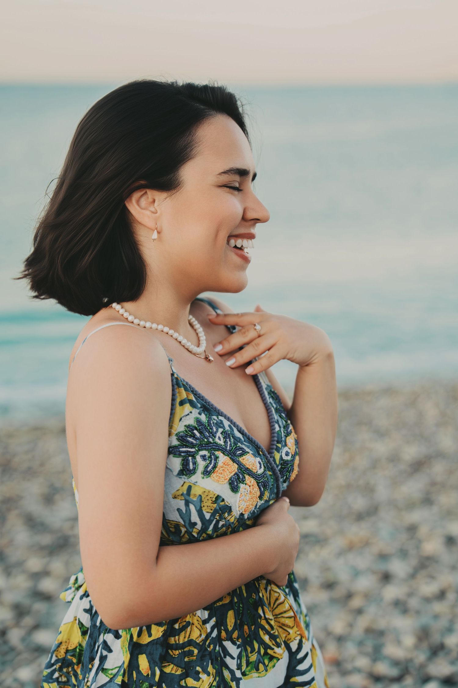 Joyful young woman in floral dress laughing on pebble beach wearing pearl necklace at golden hour.