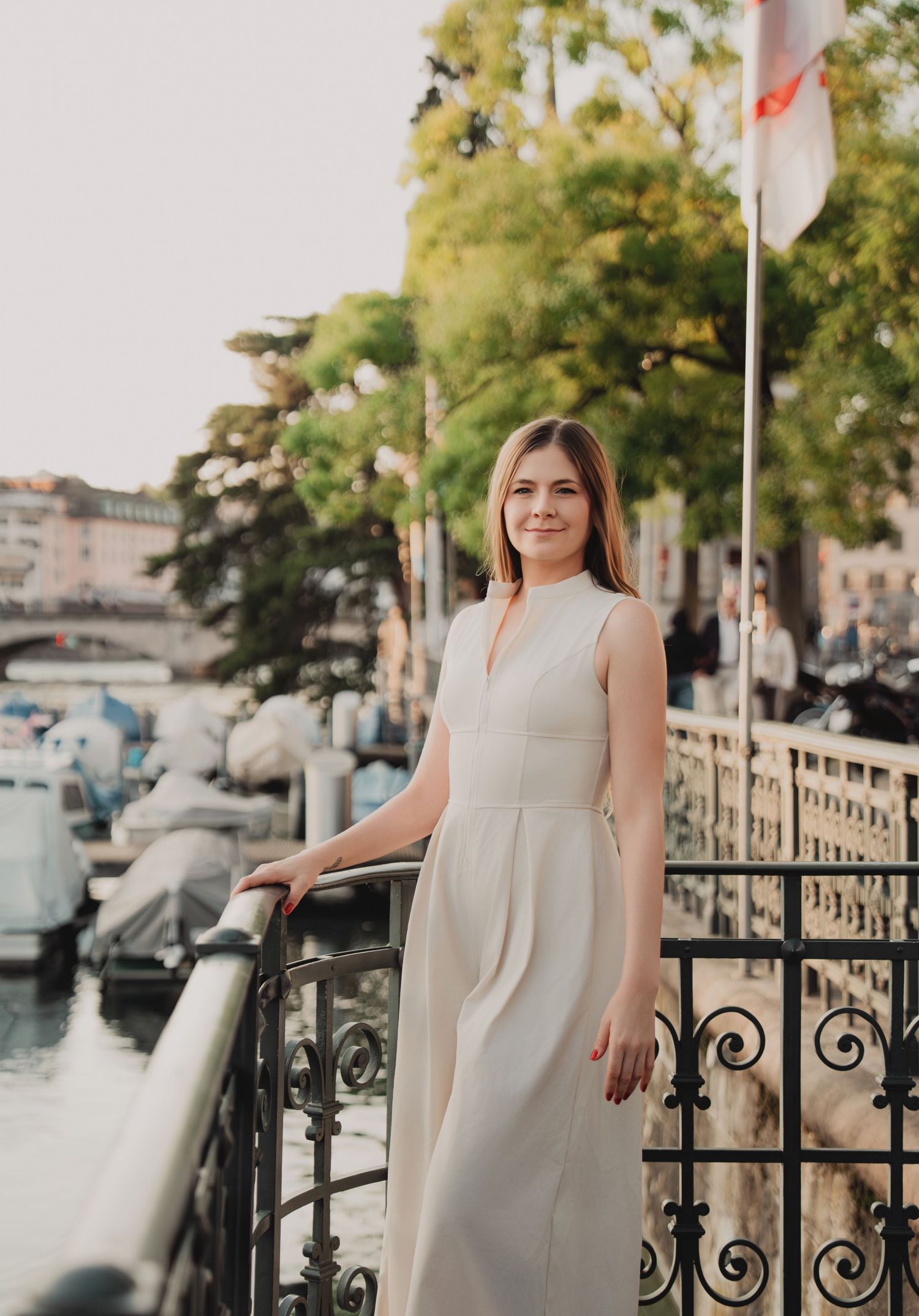 Smiling woman in white sleeveless dress standing by ornate iron railing along Zurich waterfront.