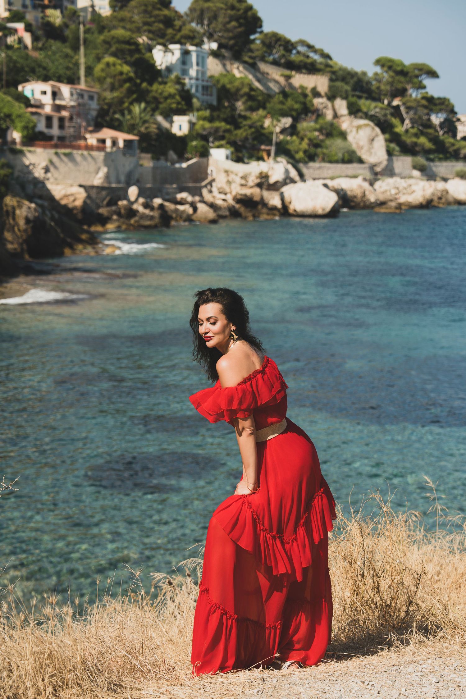 Brunette woman in red ruffled off-shoulder dress bending gracefully on Mediterranean rocky coastline.
