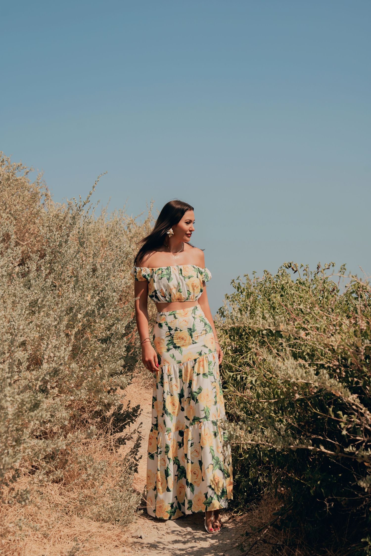 Woman in floral off-shoulder crop top and maxi skirt walking on a dry coastal path.