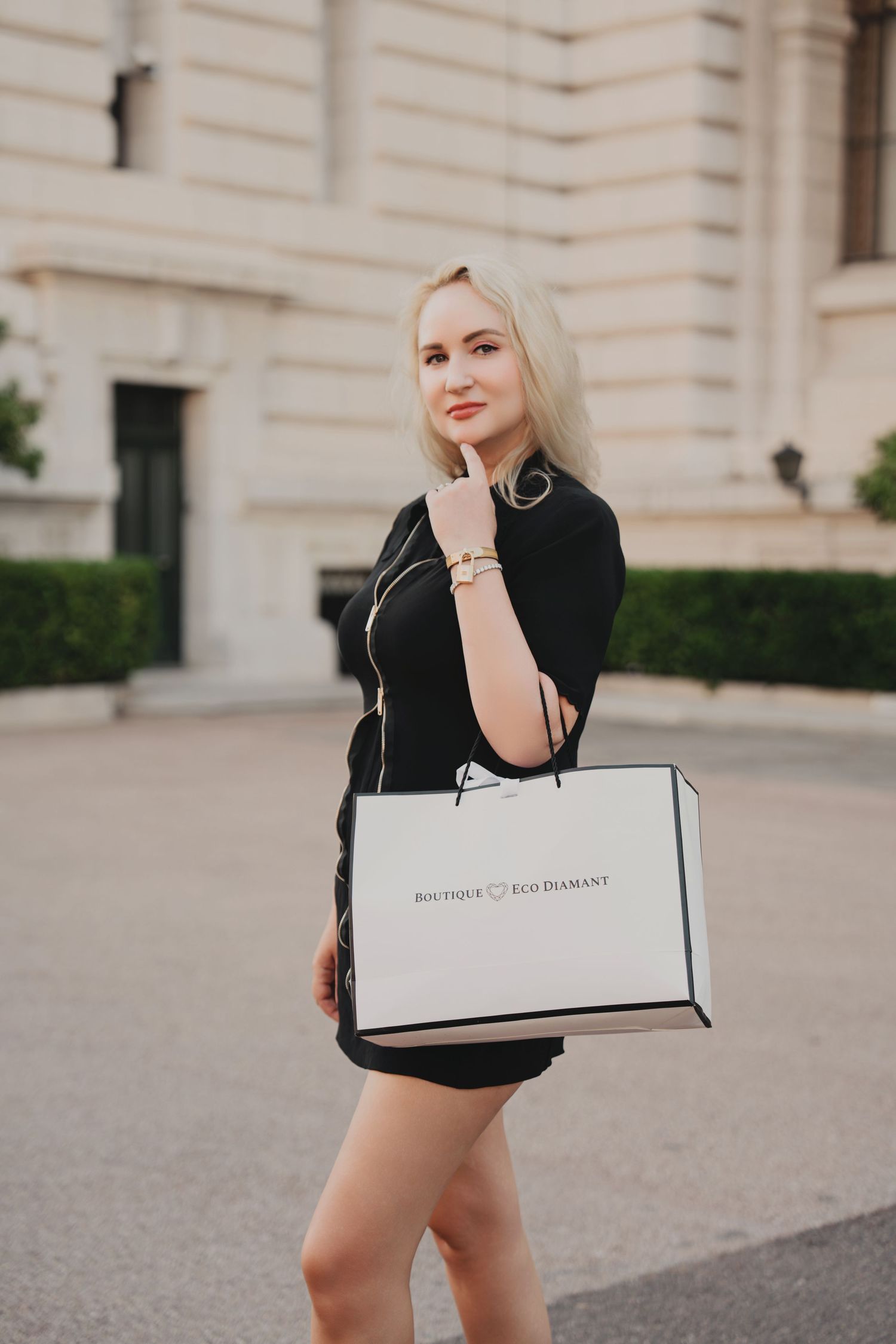 Stylish blonde woman in black dress carrying a Boutique Pur Diamant shopping bag outside a grand building.