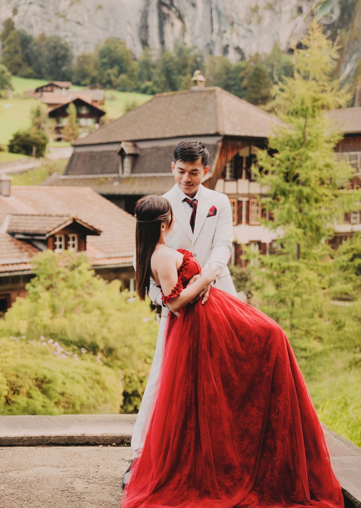Couple in red gown and white suit embracing during romantic wedding photoshoot in Swiss alpine village.