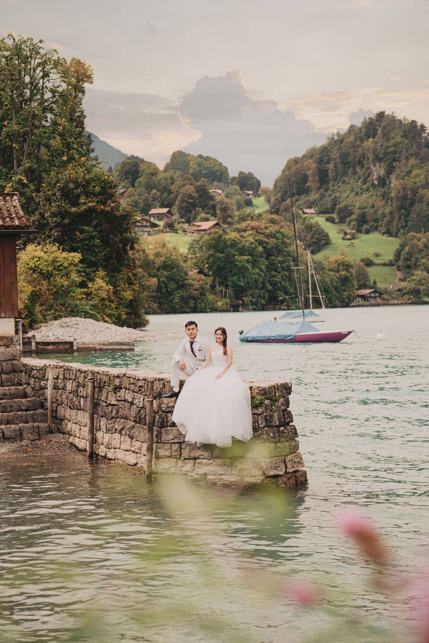 Bride and groom sitting on stone pier by alpine lake with sailboat and green hills backdrop.