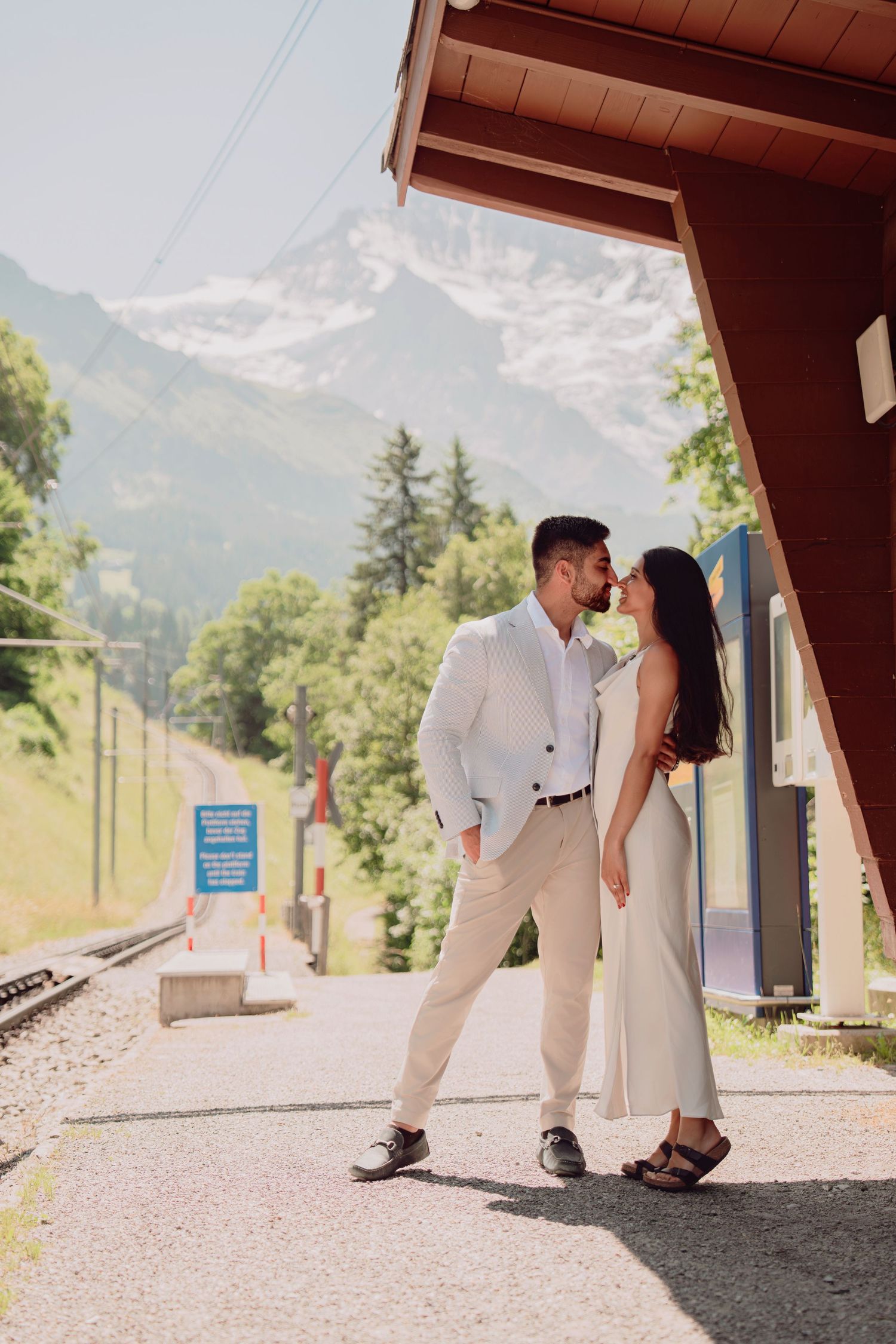 Romantic couple nearly kissing on a mountain railway platform with snow-capped Swiss Alps visible in the background.