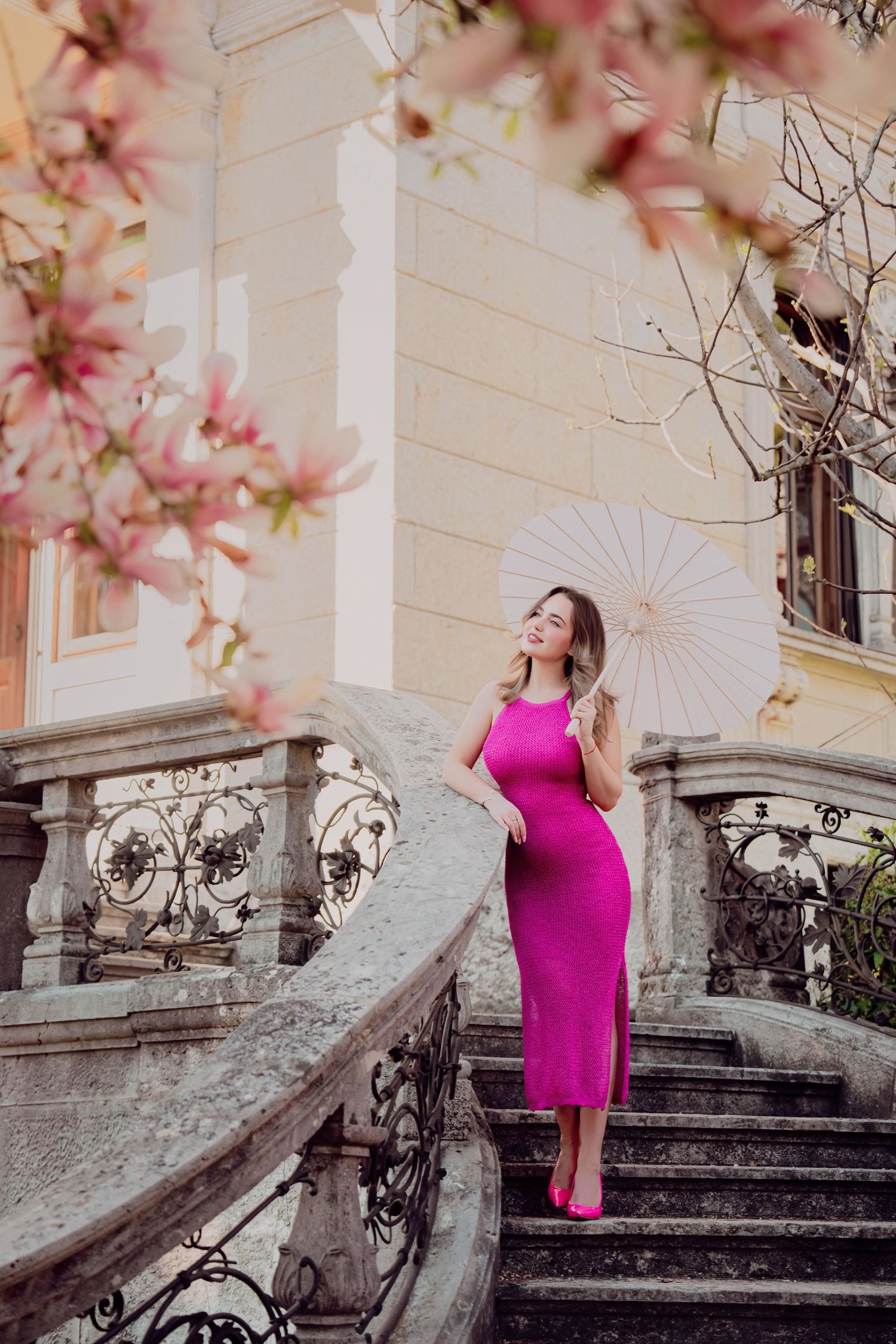 Woman in magenta dress holding parasol on elegant stone staircase with spring magnolia blossoms.