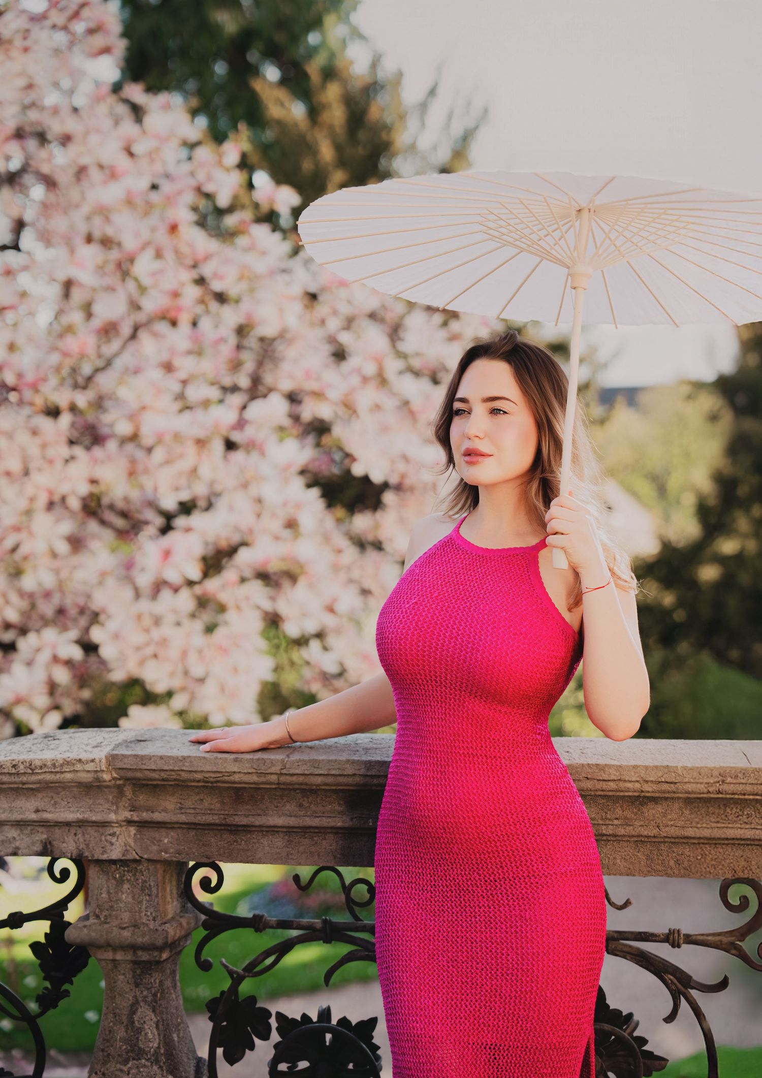 Woman in pink knit dress holding white parasol near blooming magnolia tree on ornate balcony.