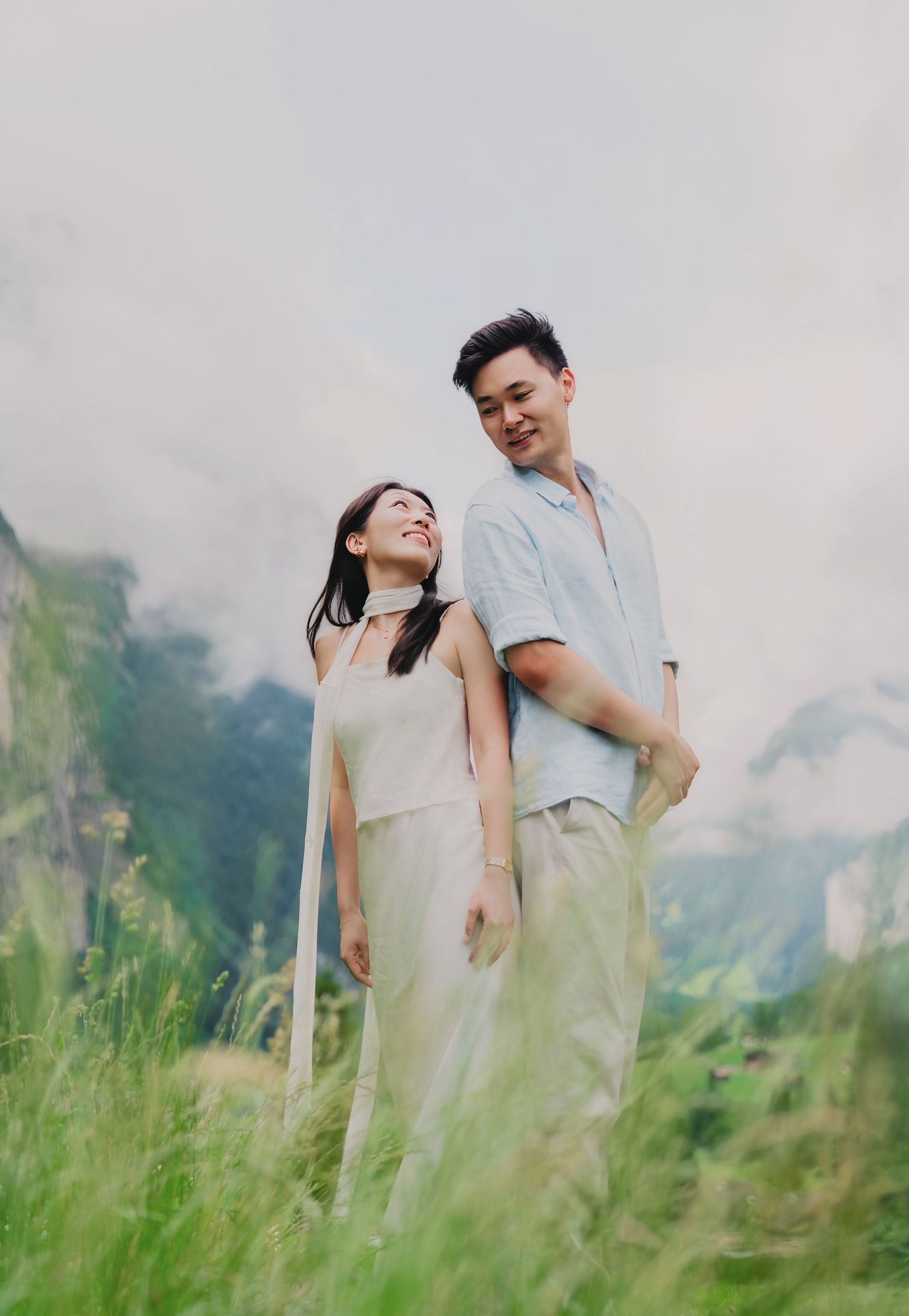 Smiling couple stands together in a misty Swiss alpine meadow, the woman looking up joyfully at her partner.