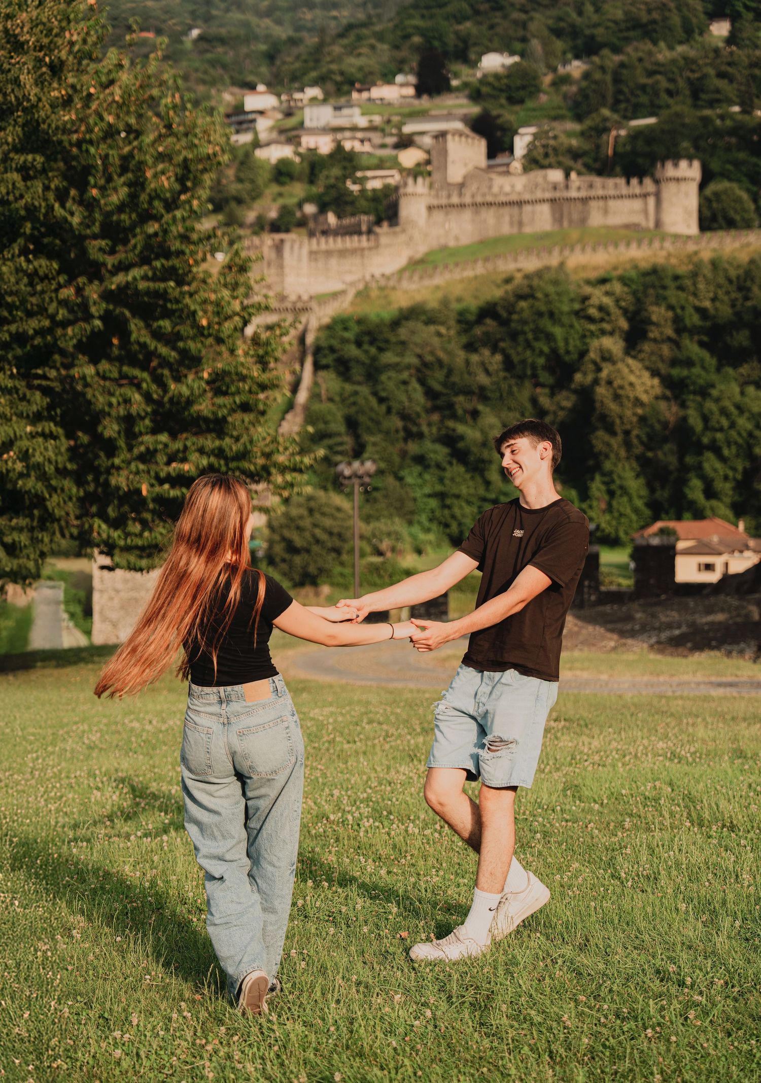 Joyful couple spinning and holding hands on a green hillside with a historic castle in the background.