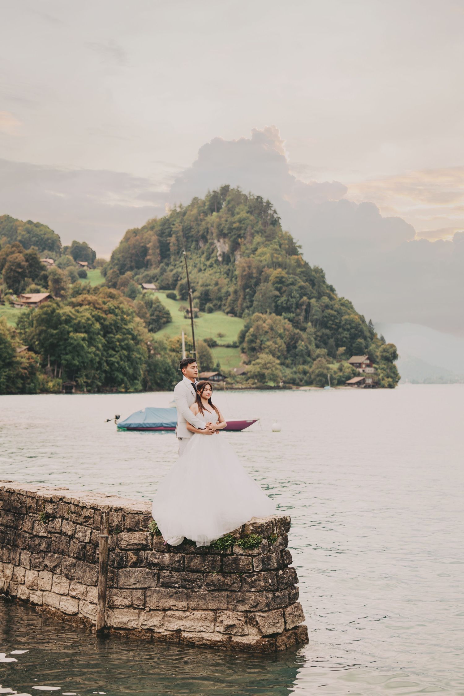 Bride and groom embracing on stone pier with sailboat and lush alpine mountain in background.