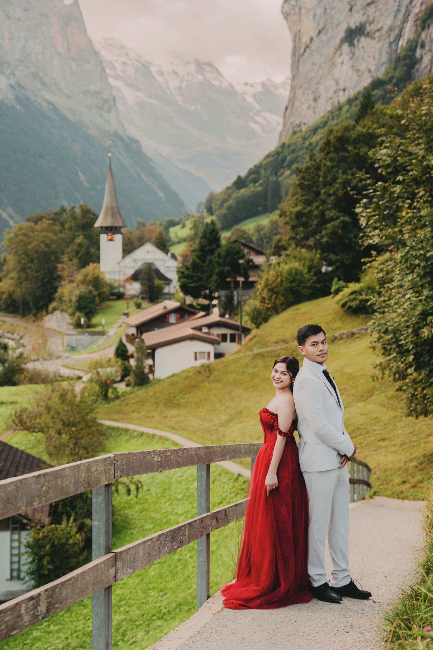 Couple in red gown and grey suit posing on a bridge with Swiss alpine village and mountains backdrop.