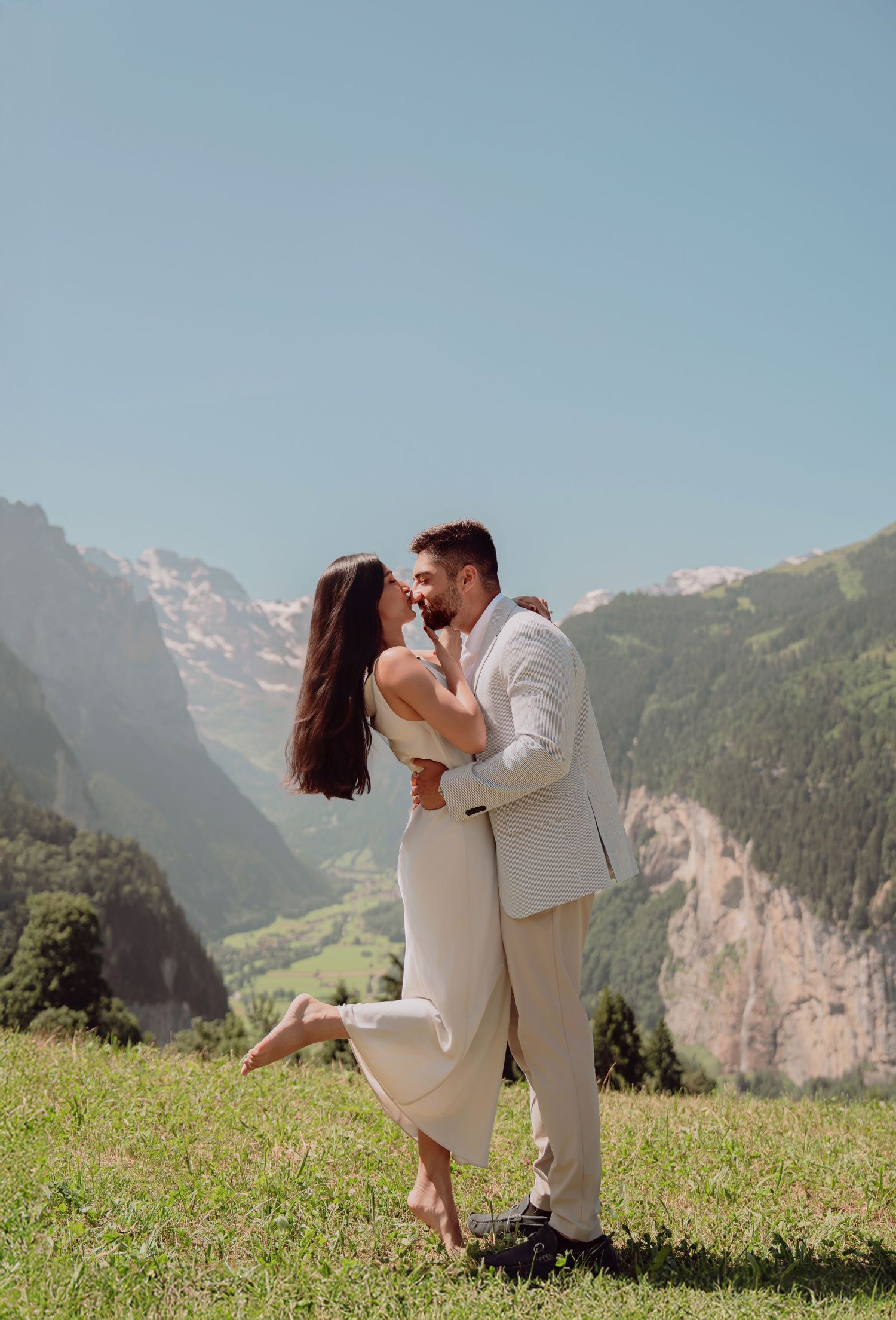 Couple sharing a romantic moment on a green hillside with stunning Swiss Alps valley backdrop on a sunny day.