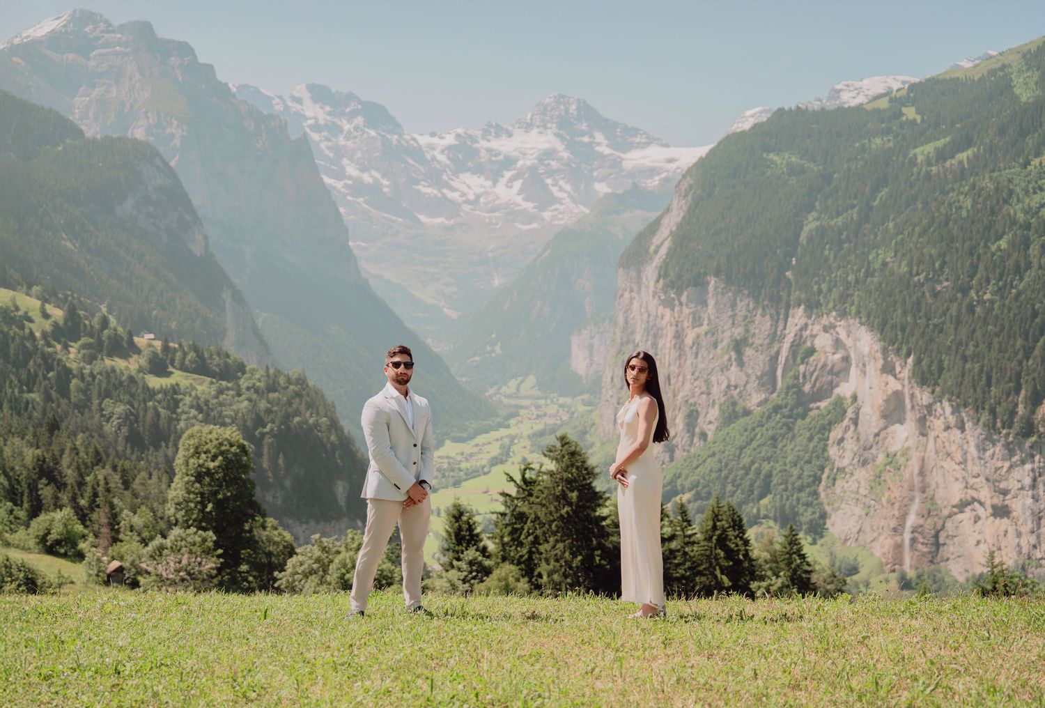 Couple posing apart on a green alpine meadow with a dramatic Swiss mountain valley stretching behind them.