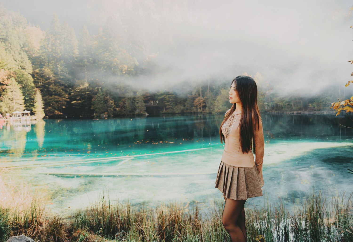 Woman in gray skirt gazes at a stunning turquoise lake with misty mountains and autumn trees.