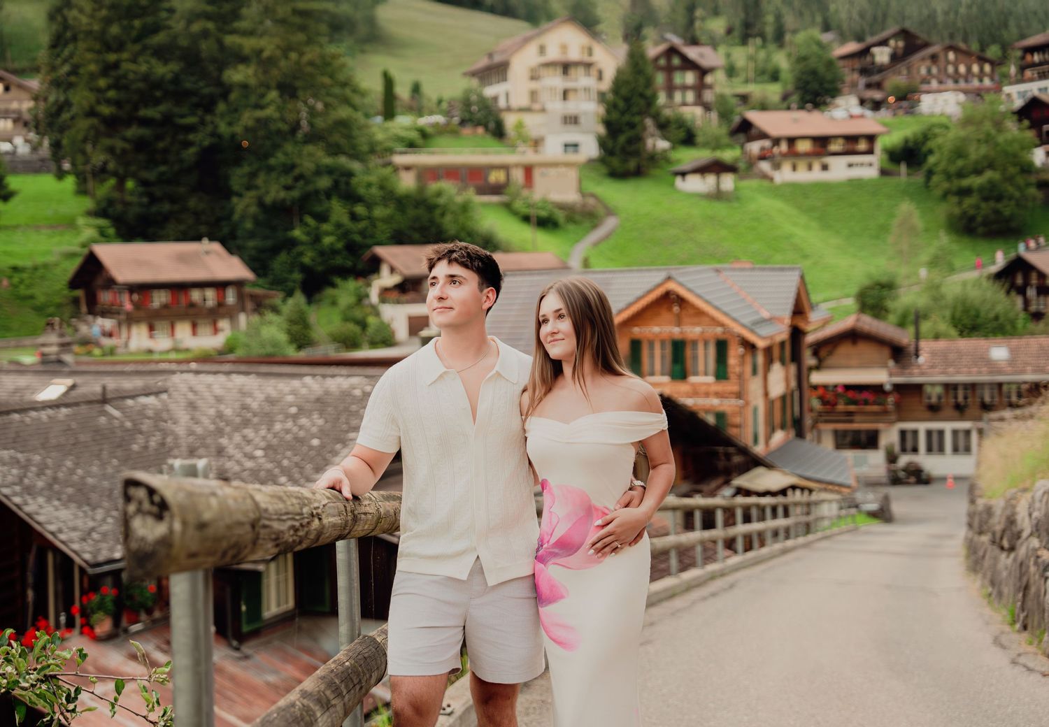 Couple leaning on wooden railing overlooking scenic Swiss village with lush green hillside chalets.
