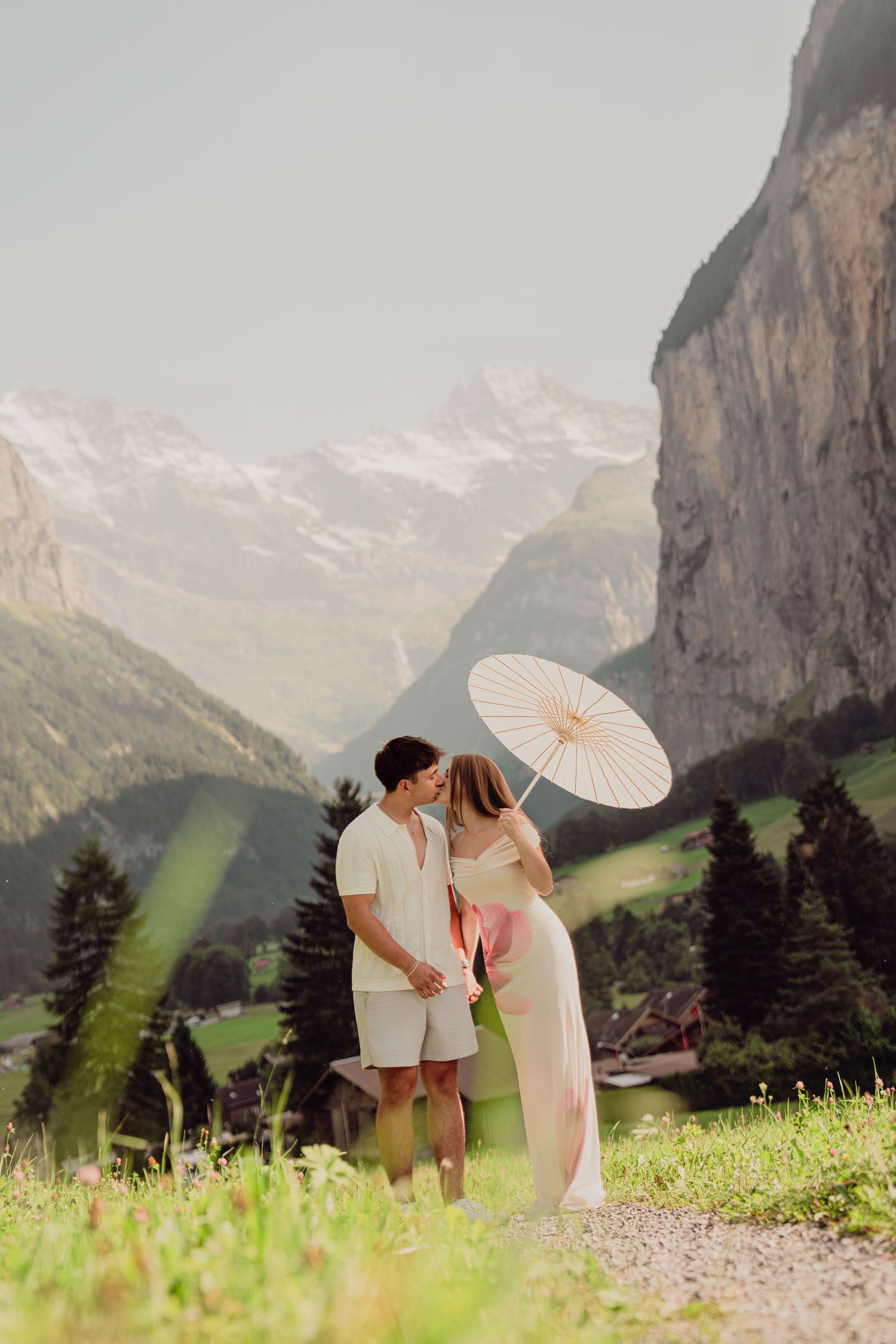 Couple kissing with white parasol on sunlit Swiss mountain path with snow-capped peaks behind.