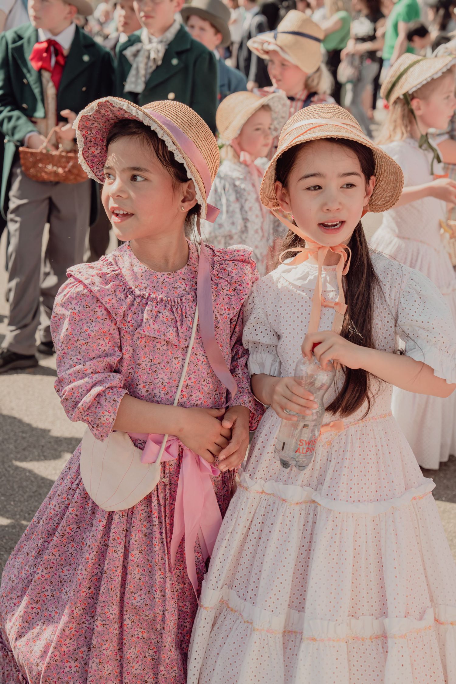 Two girls in vintage floral dresses and straw hats stand together at a traditional cultural parade.