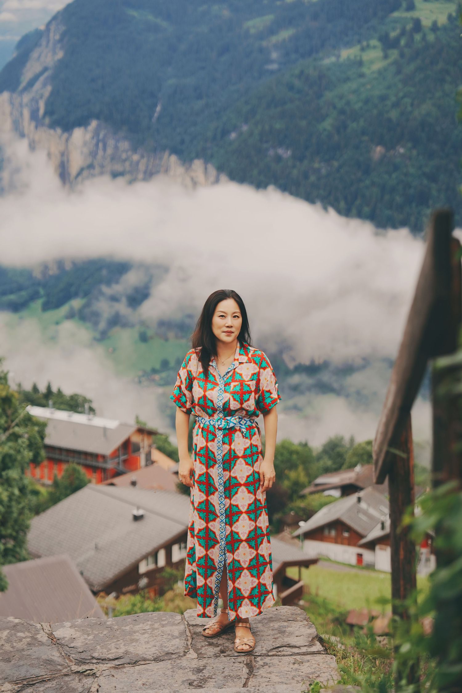 Woman in colorful patterned maxi dress stands on stone steps with misty Swiss Alpine village backdrop.
