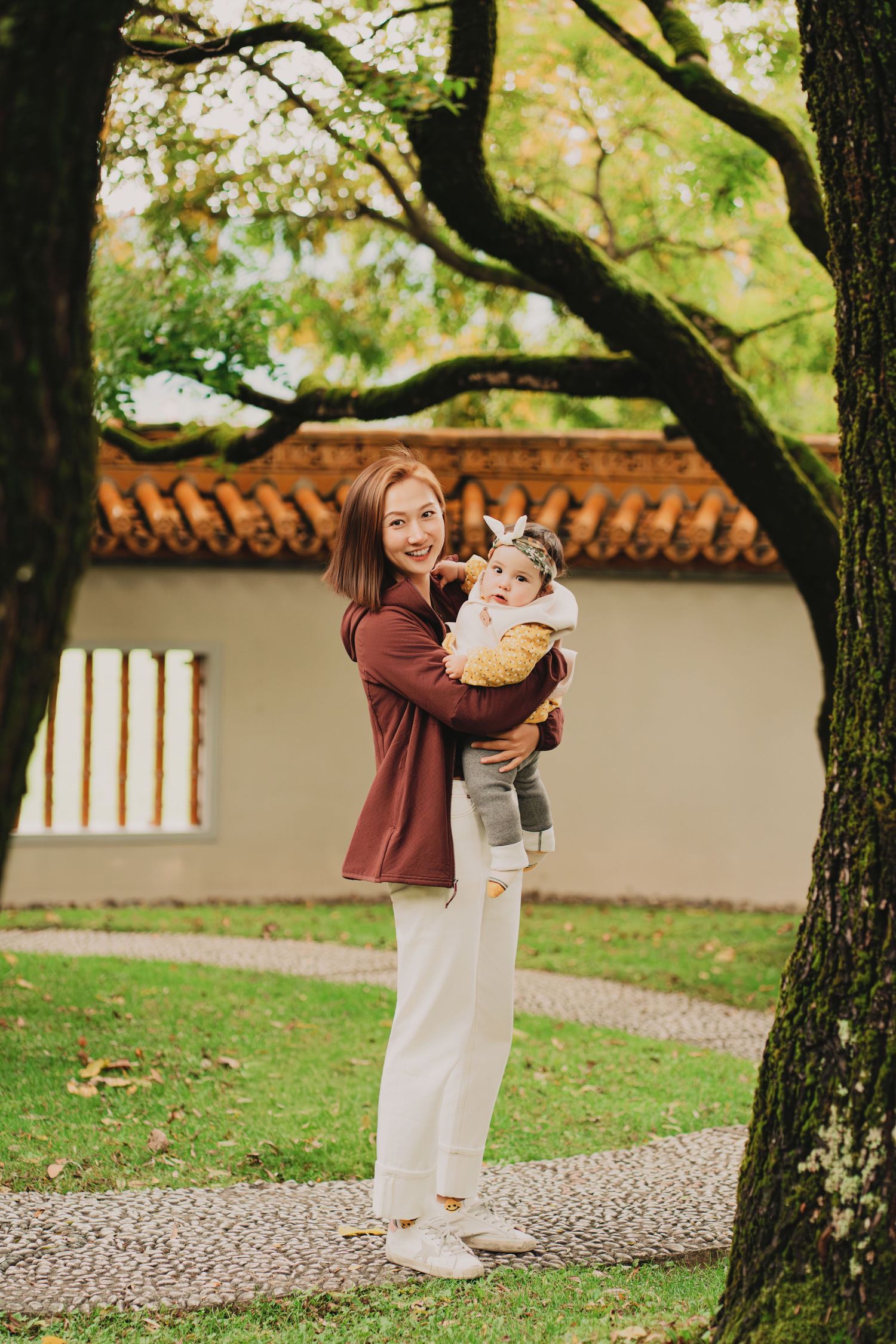Smiling mother holds baby wearing headband while standing on garden path among tall trees.