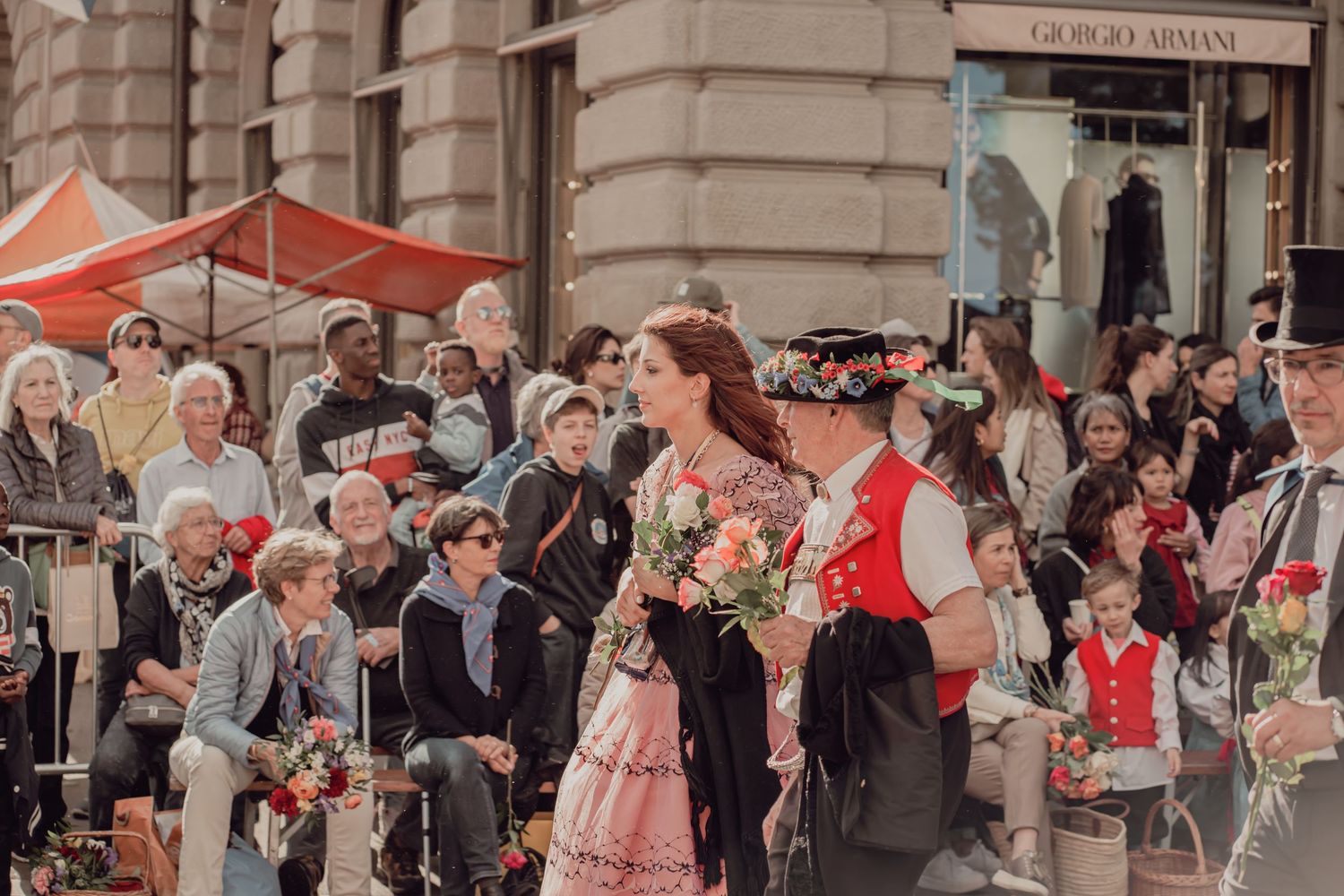Couple in traditional Swiss folk costumes walking in a street parade with spectators and Giorgio Armani store in background.