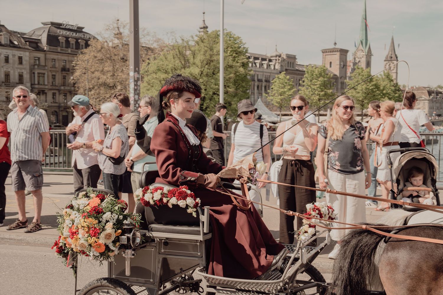 Woman in Victorian burgundy dress riding decorated horse carriage through Zurich streets.
