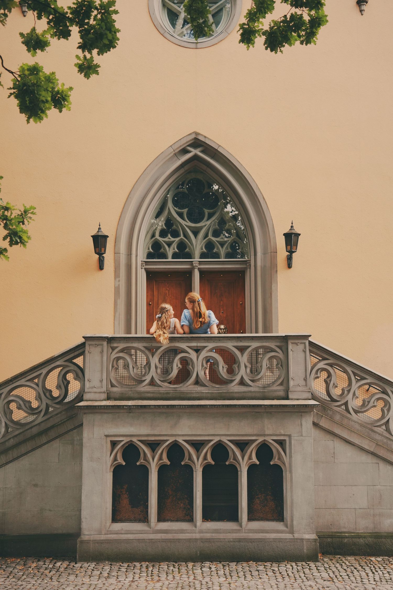 Two girls stand on an ornate Gothic stone balcony of a yellow European building.