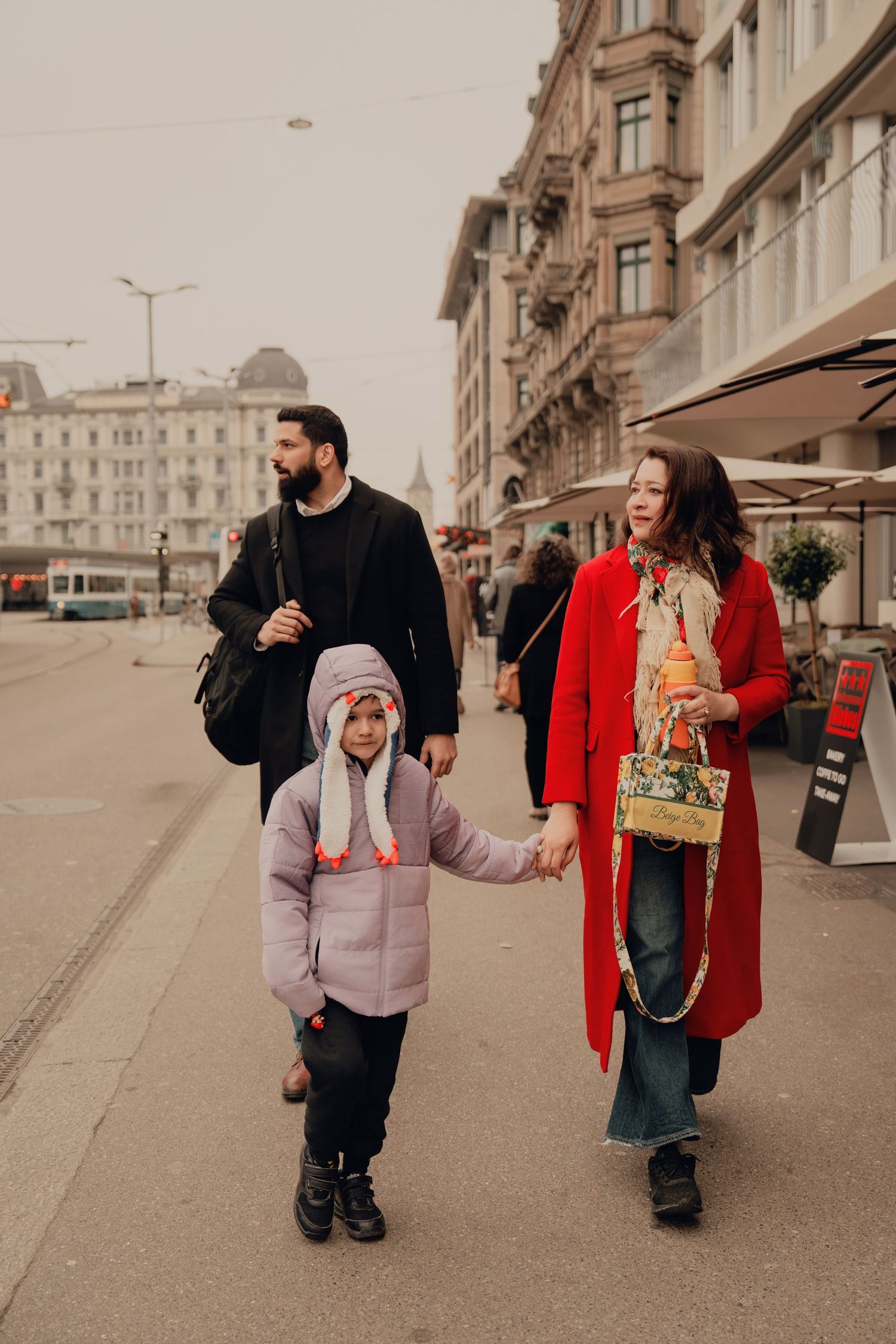 Family of three walking on European city street, woman in red coat holding young child's hand.