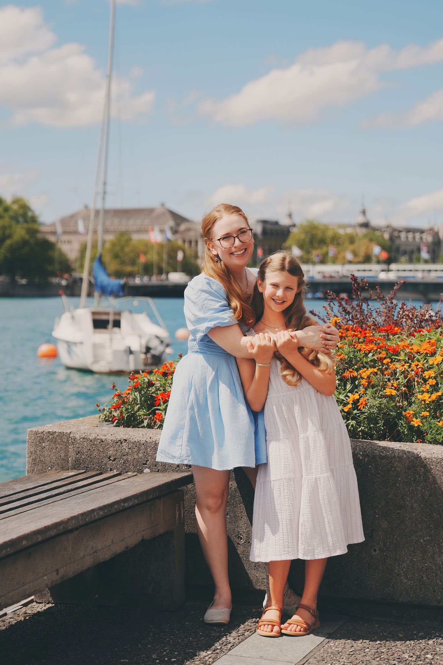 Mother hugging daughter by colorful flowers at sunny lakeside promenade with sailboats.