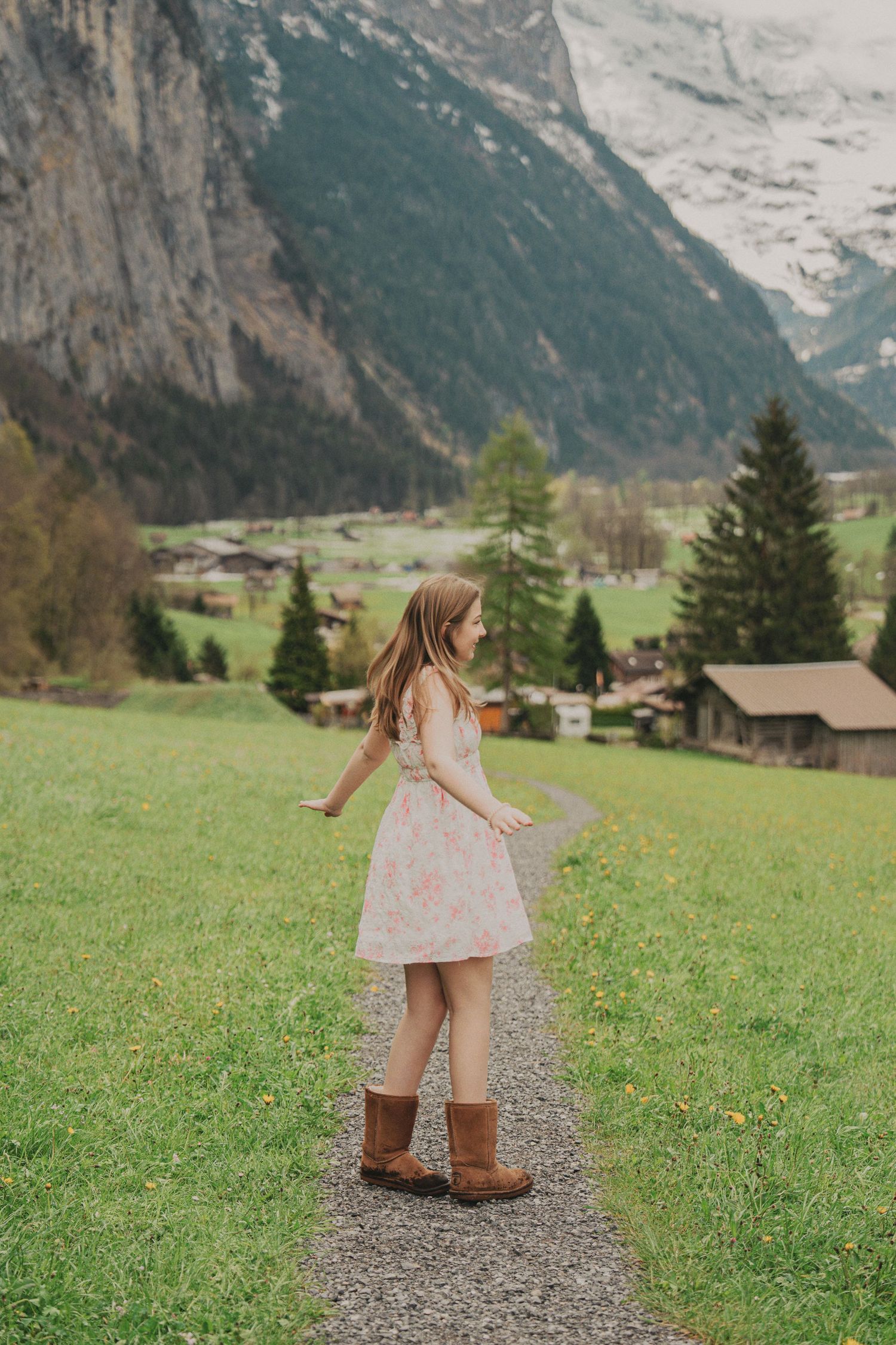 Young woman in pink floral dress and brown boots standing on a mountain path with arms outstretched in Alpine valley.