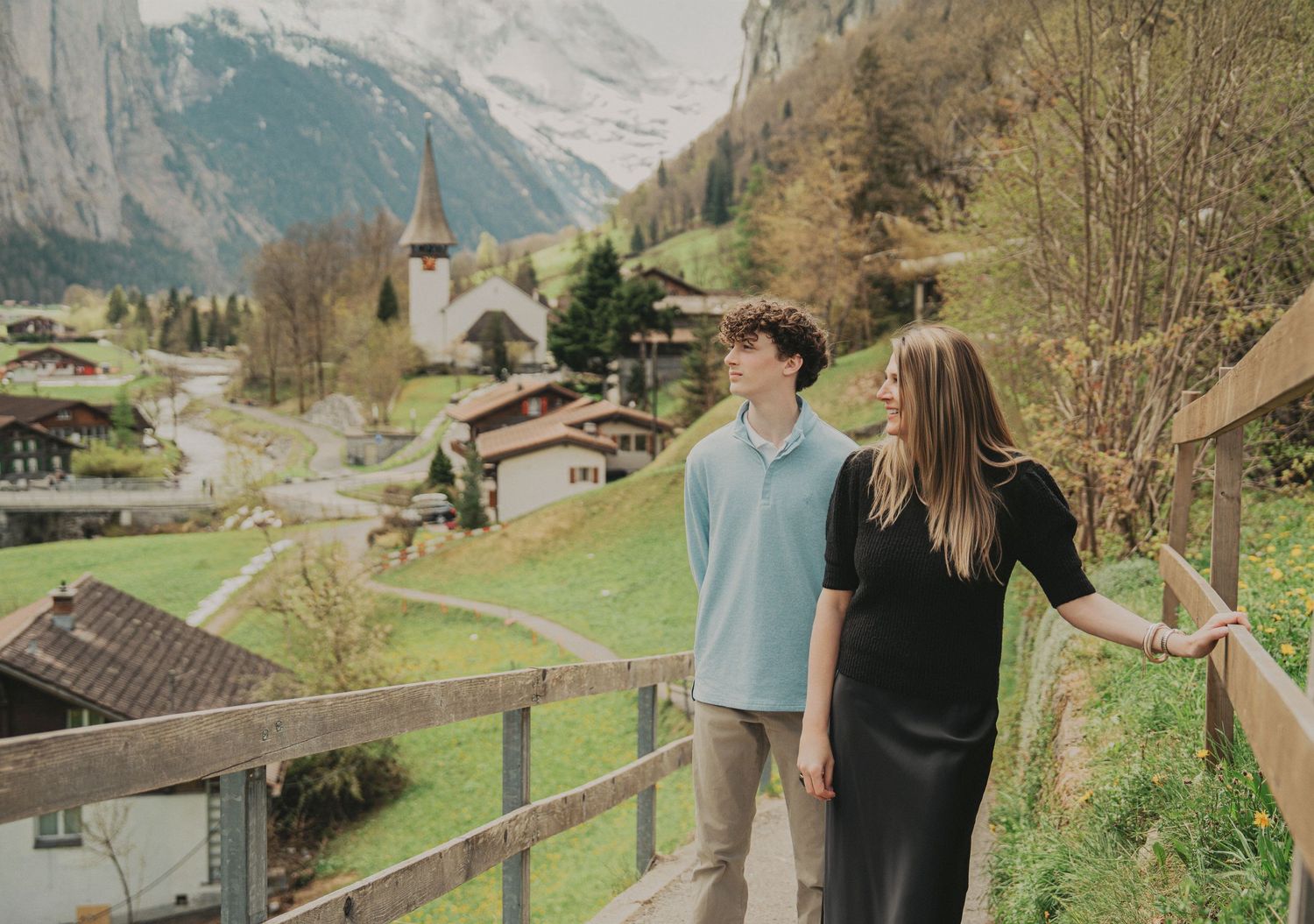 A teenage boy and woman walk together on a wooden bridge with a charming Swiss mountain village behind them.