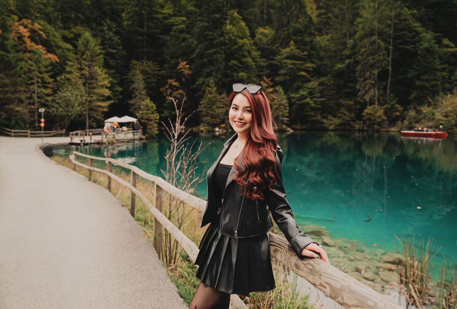 Smiling woman with long red hair in black leather outfit posing beside vibrant turquoise alpine lake.
