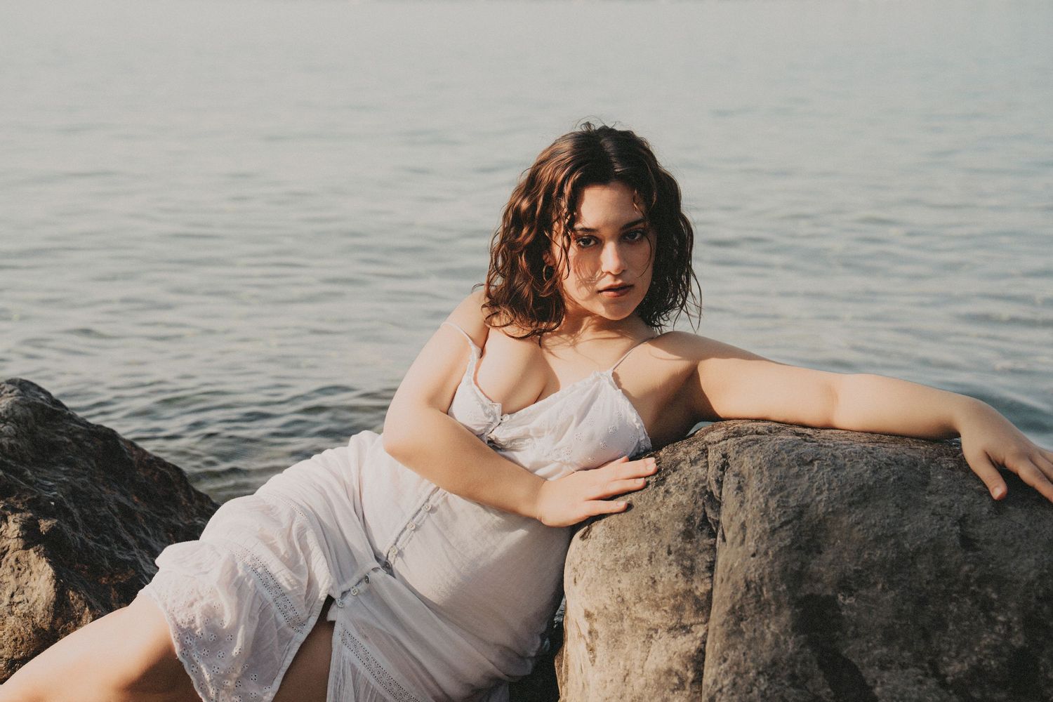 Woman in white dress reclining against large coastal rocks by calm sea.