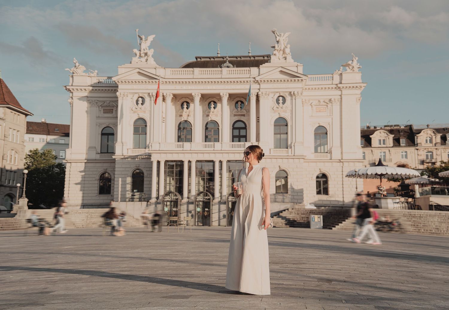 Woman in elegant white dress posing in front of grand neoclassical opera house facade.