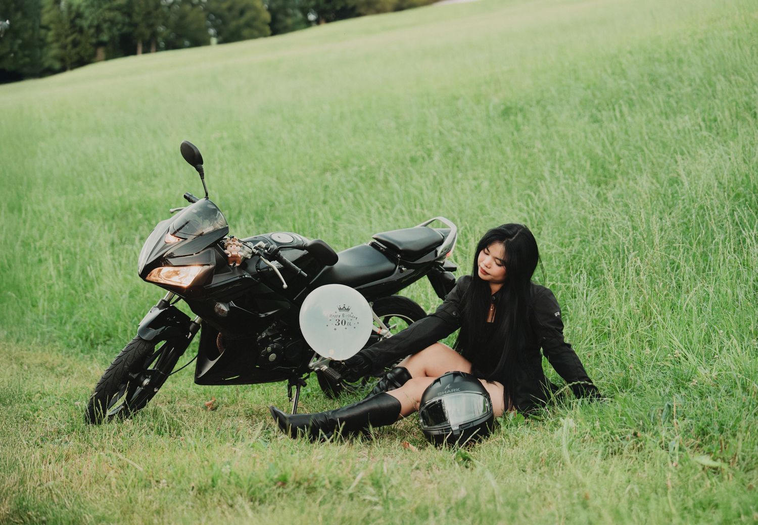 Woman in black jacket sitting on grass beside a black sport motorcycle holding a helmet.