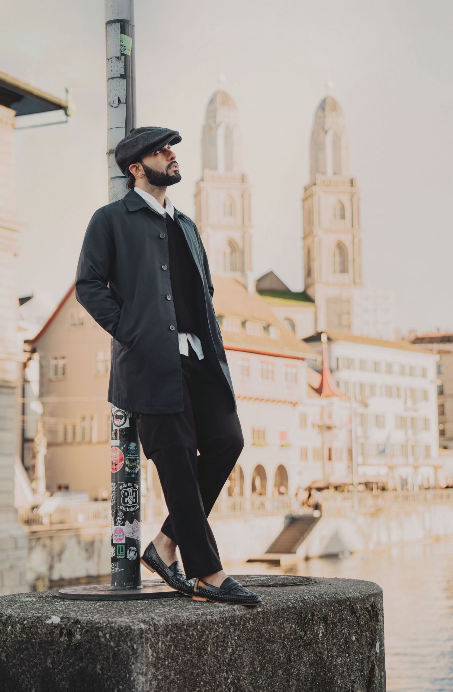 Stylish man in long dark coat and flat cap standing on a stone pillar with Grossmunster church behind.