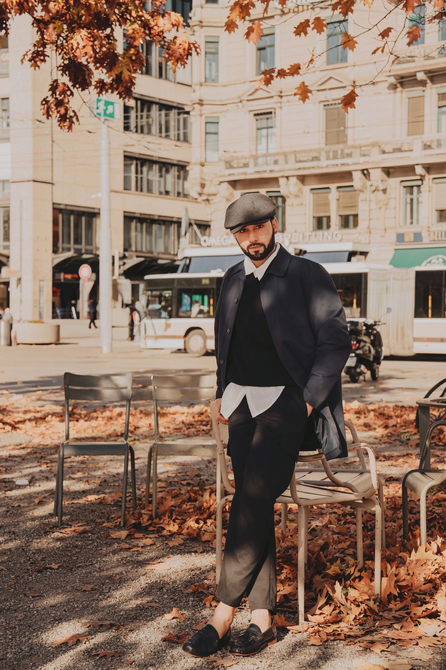 Dapper bearded man in dark coat and cap posing with autumn leaves in city square.