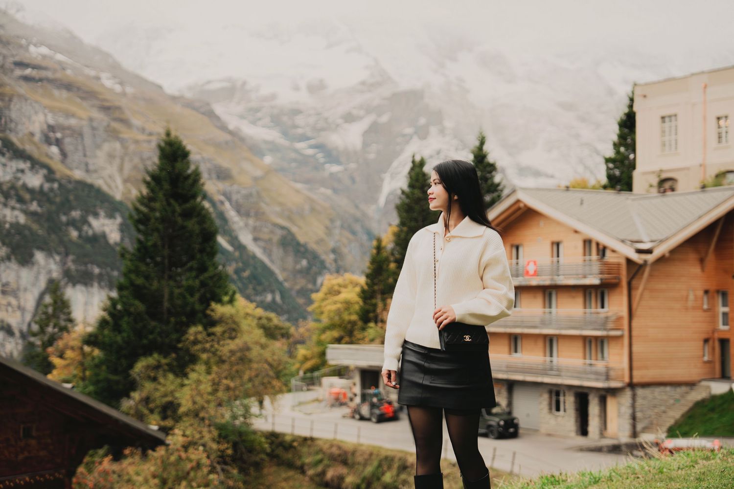 Woman in white sweater and black skirt stands before misty Swiss mountain village scenery.