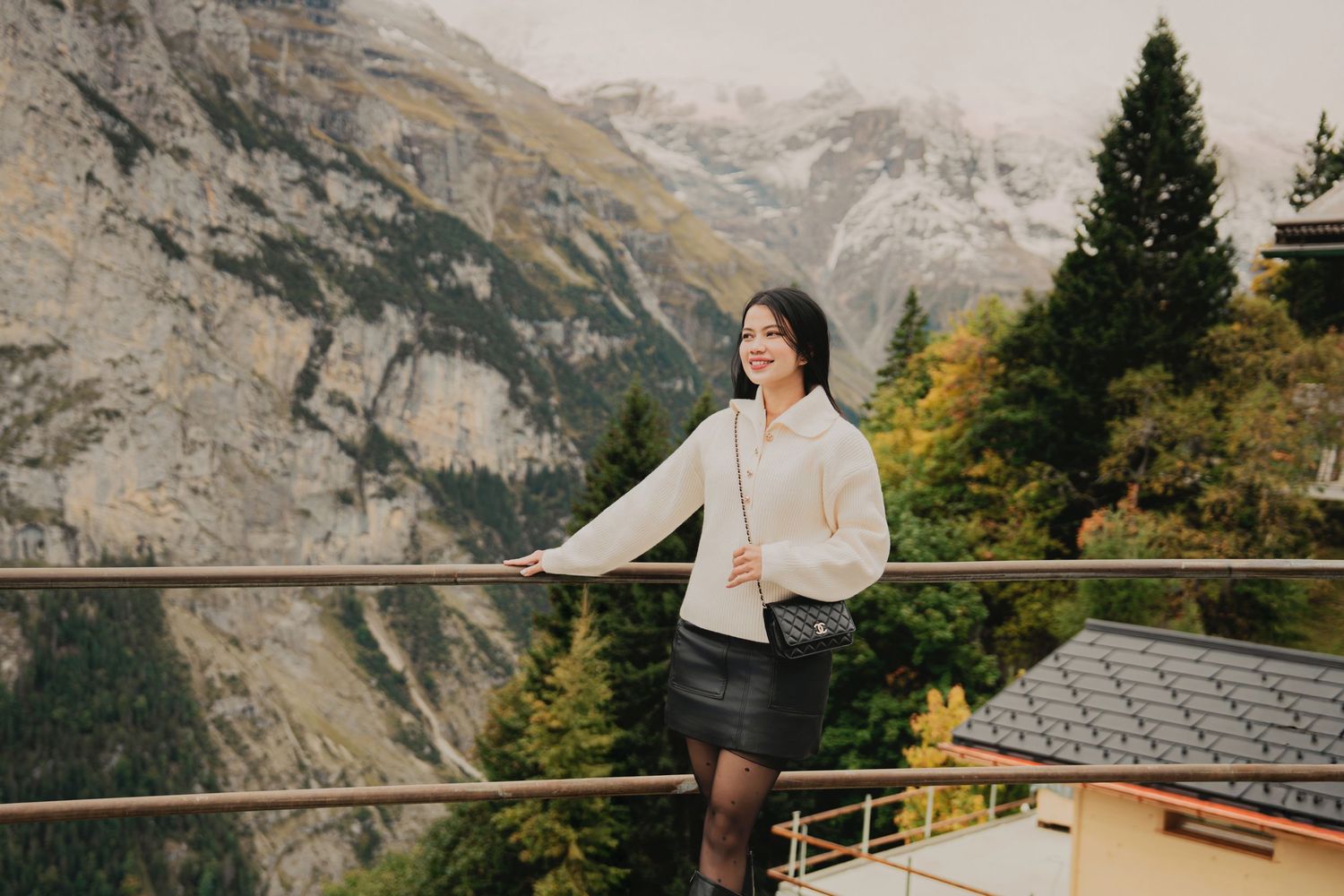 Stylish woman in white sweater leans on railing with dramatic Swiss Alps mountain scenery behind her.