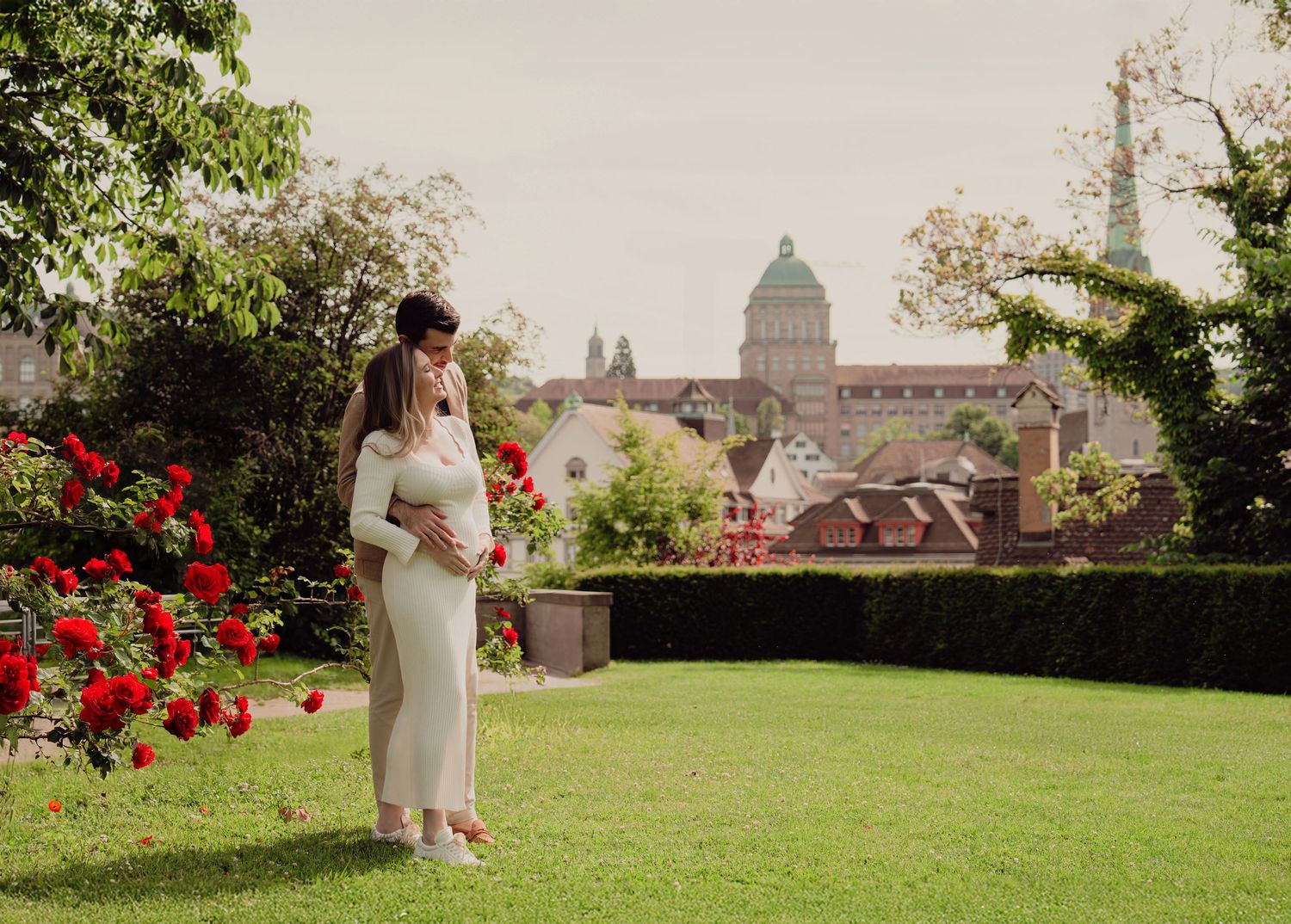 Pregnant woman and partner embracing in a garden with red roses and European cathedral skyline.