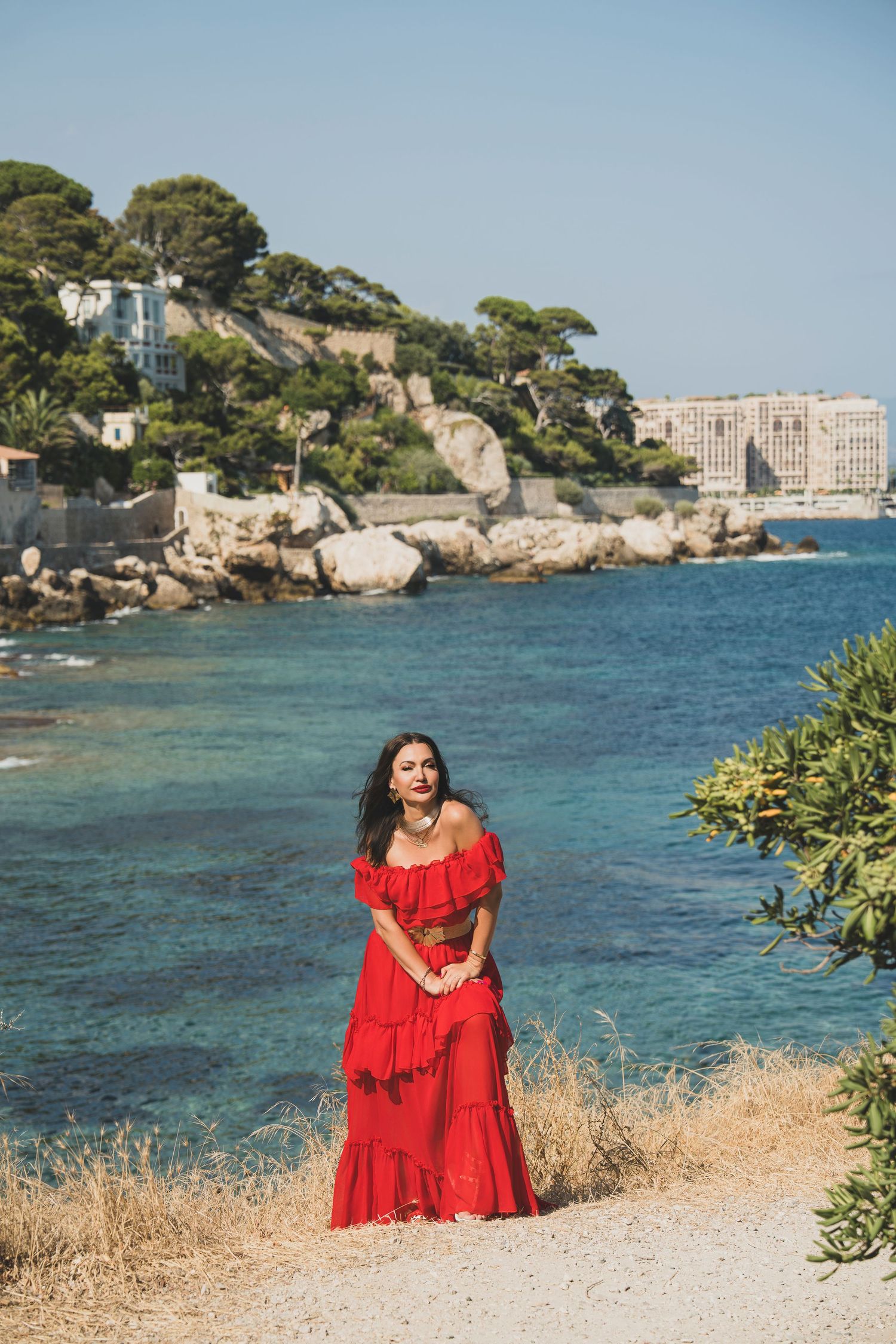 Woman in red ruffled off-shoulder dress standing on rocky coastline with turquoise sea backdrop.