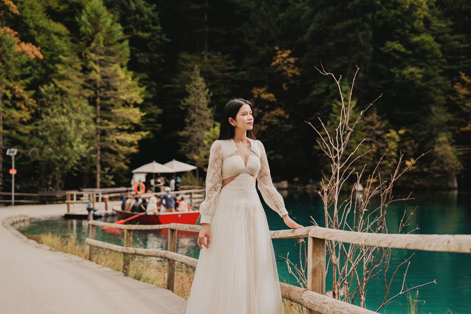 A woman in an elegant white lace dress stands by a turquoise alpine lake surrounded by forest.