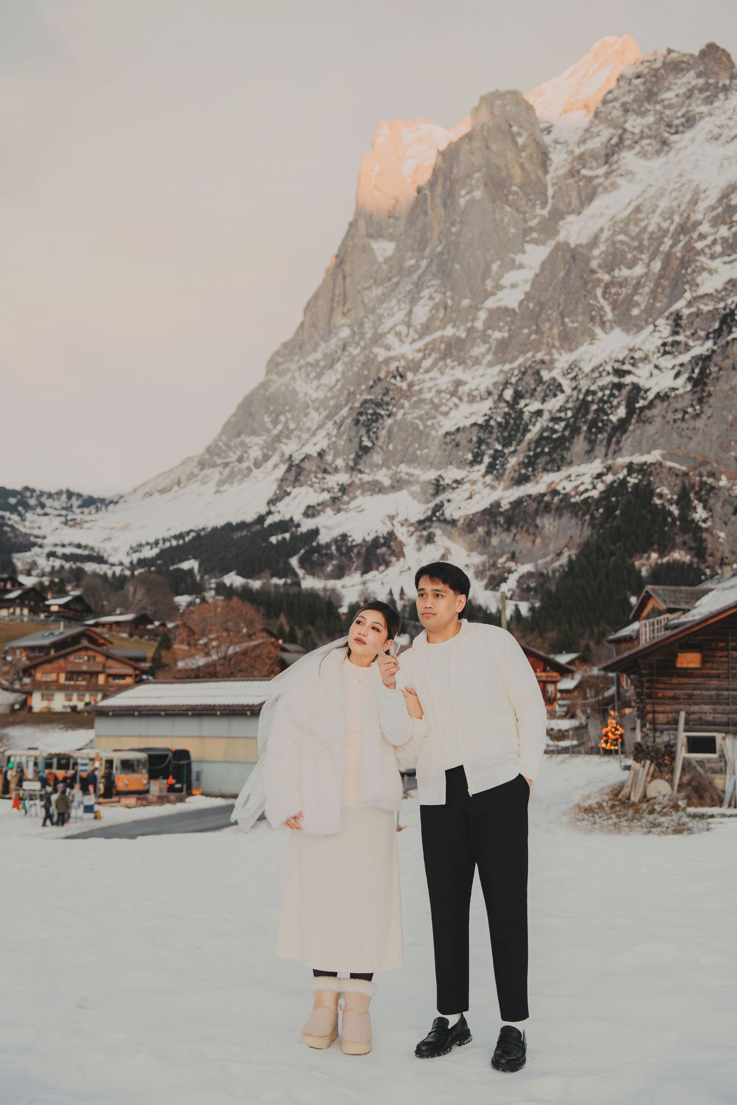 Couple posing in white winter clothing in snowy Grindelwald village with illuminated mountain peak at dusk.