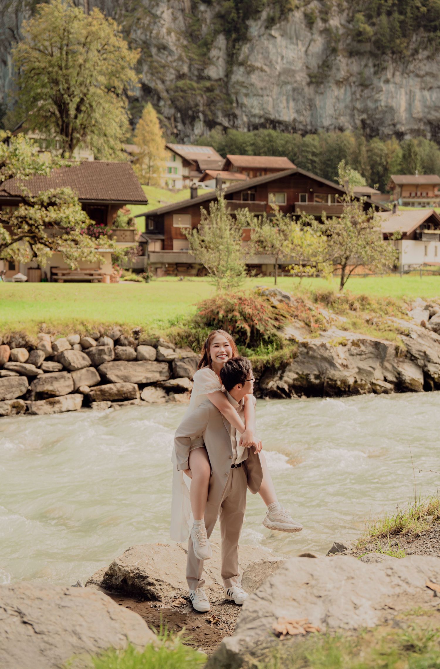 Man carrying woman on his back on rocks beside a turquoise river in a scenic Swiss alpine village.
