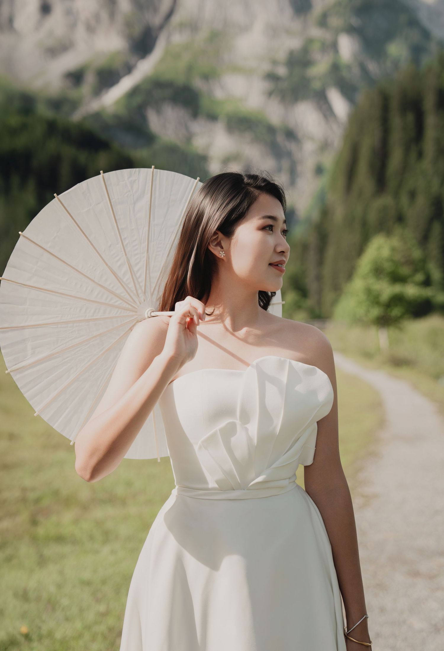 Bride in strapless white gown holding a white paper parasol with Alpine mountain scenery behind.