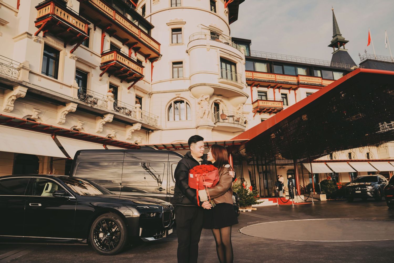 Asian couple embracing with red roses in front of a grand luxury hotel with black cars.