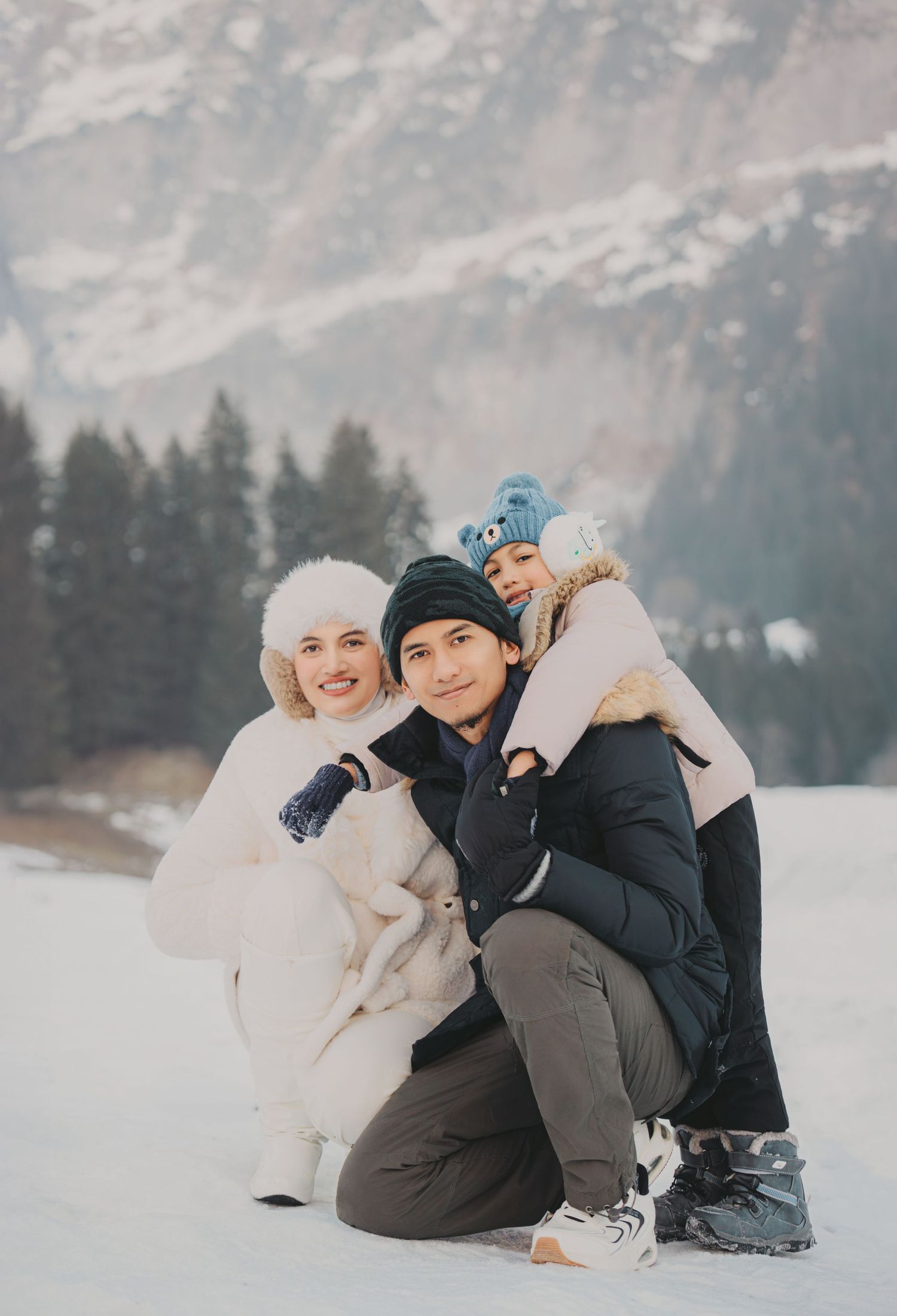 Happy family of three posing together in snowy alpine landscape with mountains and pine trees in background.